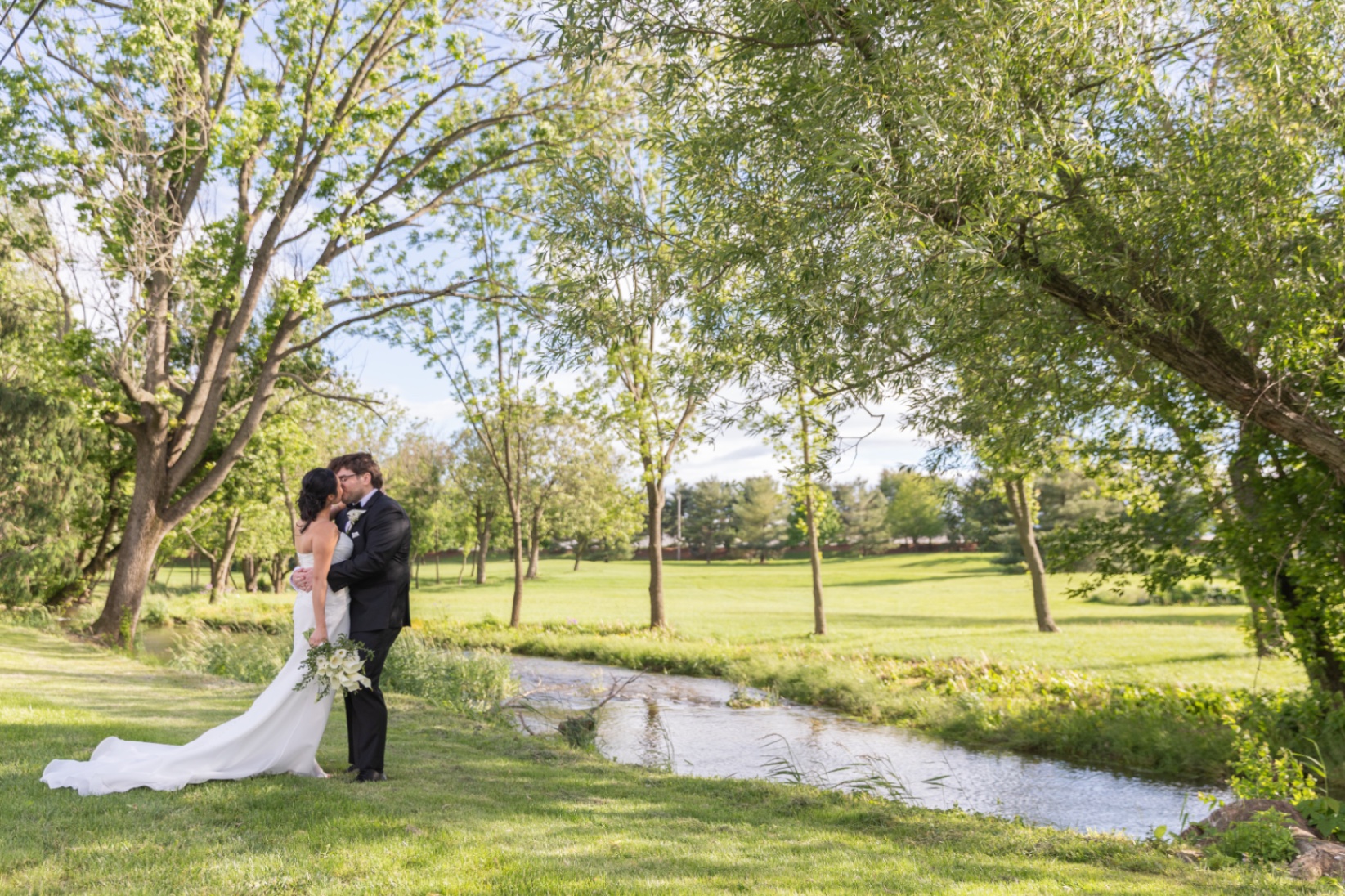A bride and groom share a kiss by a gentle creek in a sunlit park