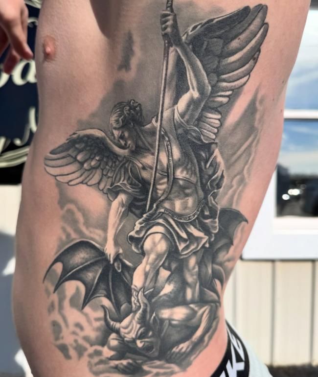 Detailed black and grey tattoo of Saint Michael, showcasing dynamic shading and intricate armor detail across the side ribs.