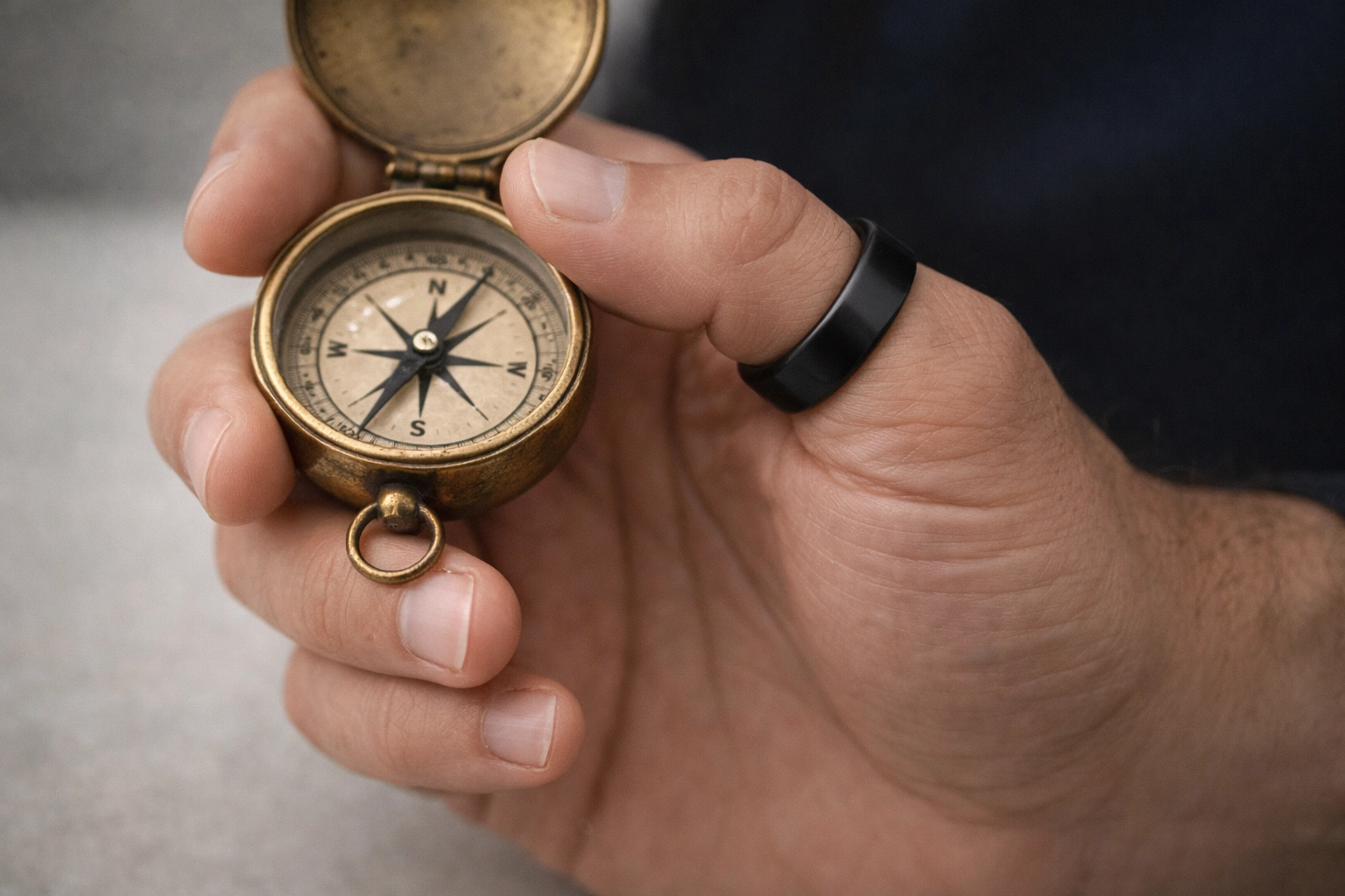 Man’s hand featuring a black thumb ring and a cobalt index finger ring holding a vintage compass.