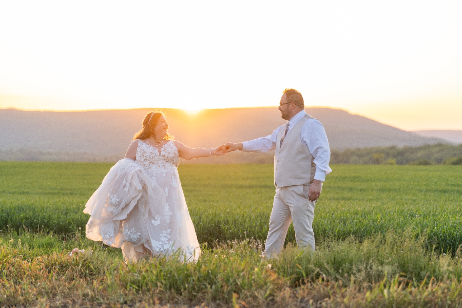 Bride and groom holding hands and smiling at each other in a sunlit field at sunset