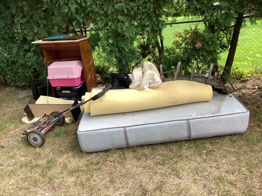 Assorted junk items staged in a backyard, including an old mattress, foam padding, and worn-out furniture ready for removal
