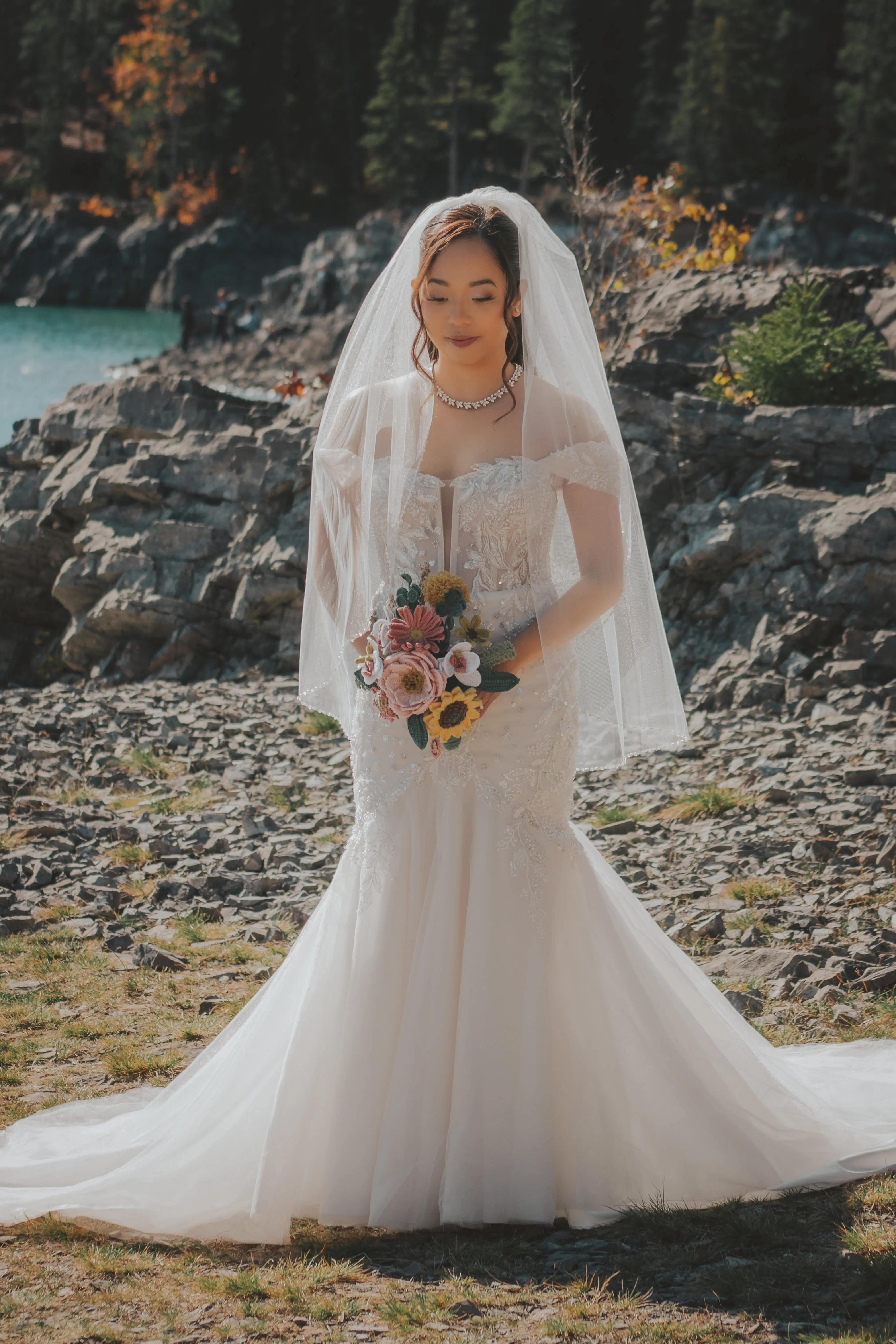 bride-lace-mermaid-dress-turquoise-lake-banff.webp A bride in a lace mermaid wedding dress and veil stands beside a turquoise mountain lake, holding a pastel bouquet. The rocky shoreline and evergreen trees provide a scenic backdrop, showcasing the natural beauty of Banff's elopement locations.