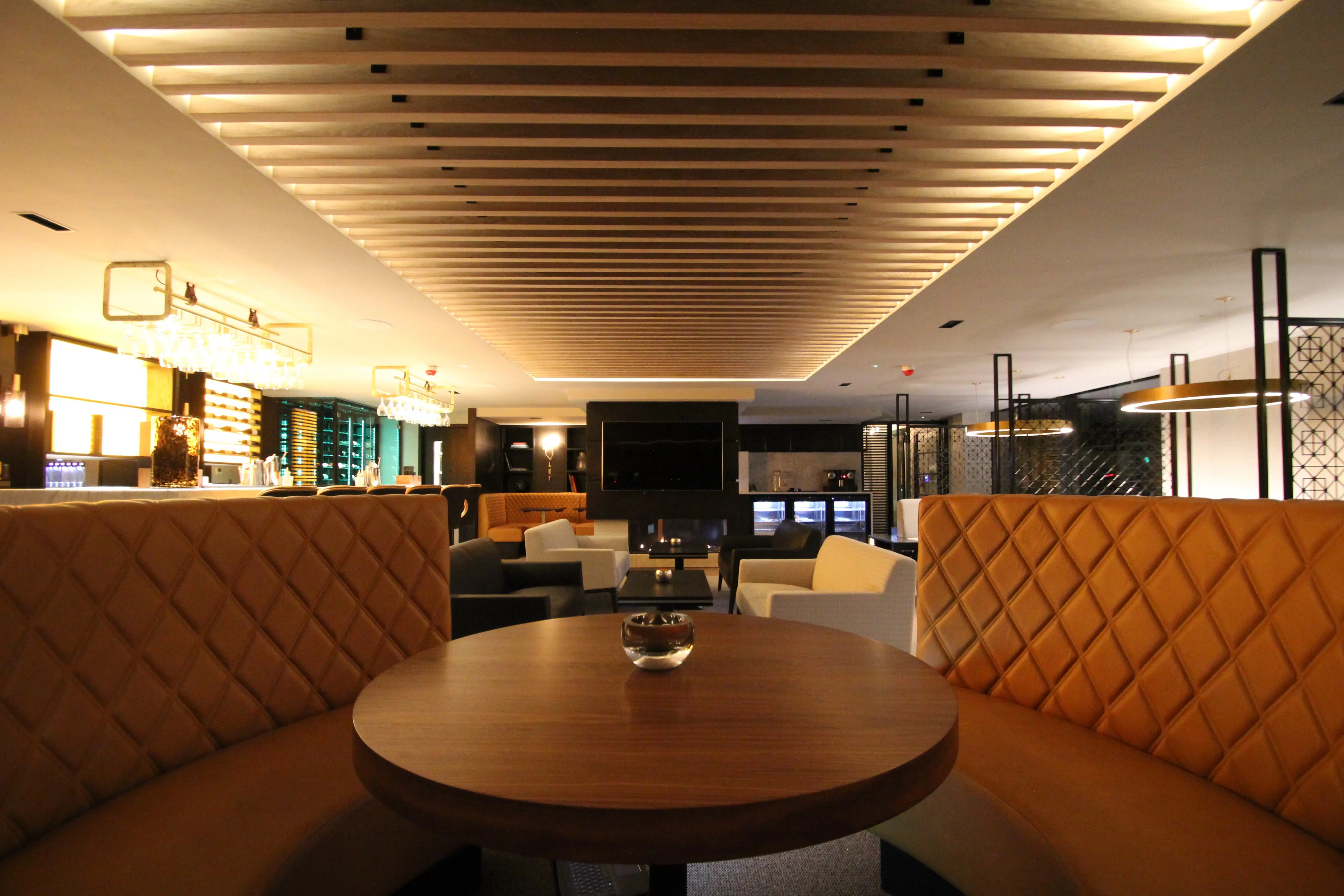 Stylish lounge area featuring luxurious quilted booths, a modern wooden ceiling, and elegant bar and kitchen facilities.