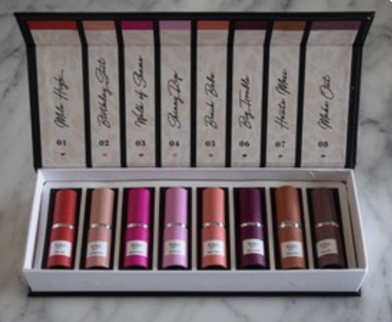 Eight Hickey Lipstick refillable pendant-style cases arranged in a marble-patterned box, including the 'Big Trouble' shade. Clean, high-contrast flat lay with a modern editorial feel.