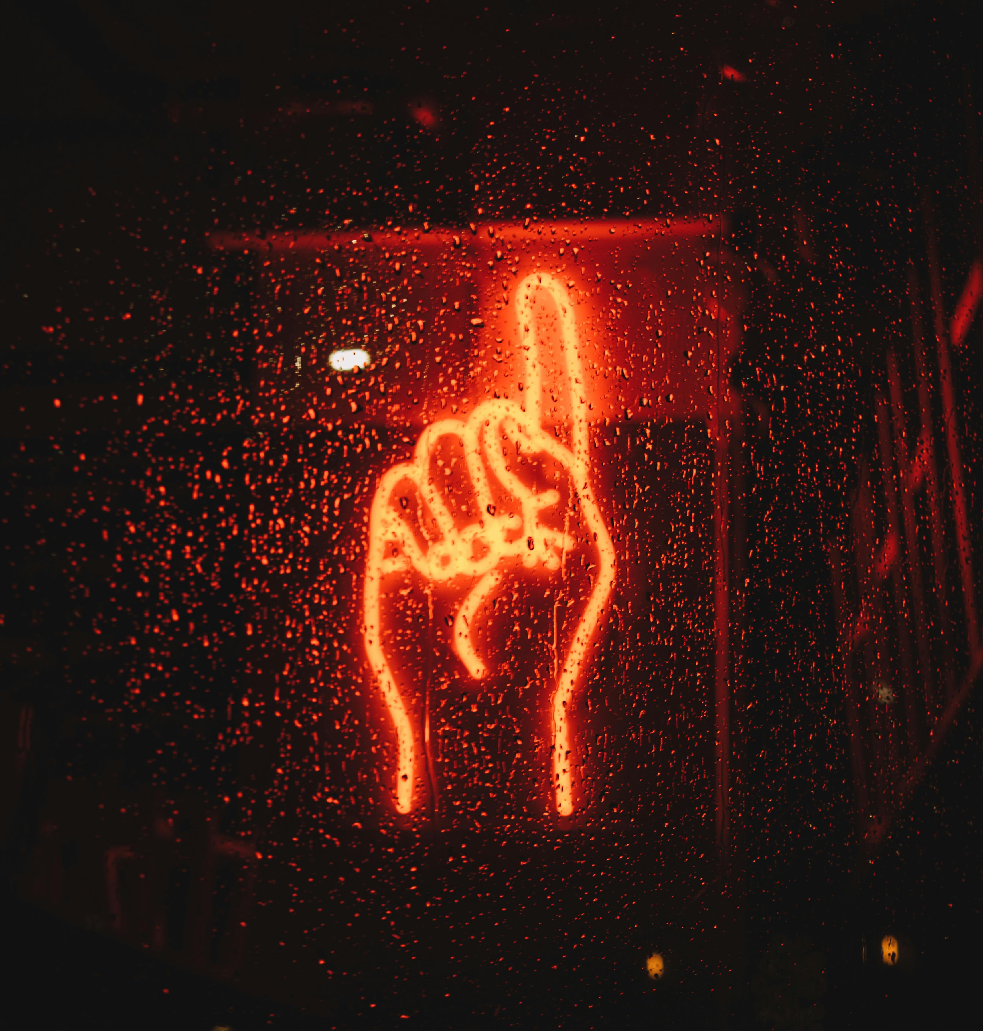 Red LED neon sign shaped as a hand with the index finger pointing upward, glowing vividly behind a rain-speckled glass