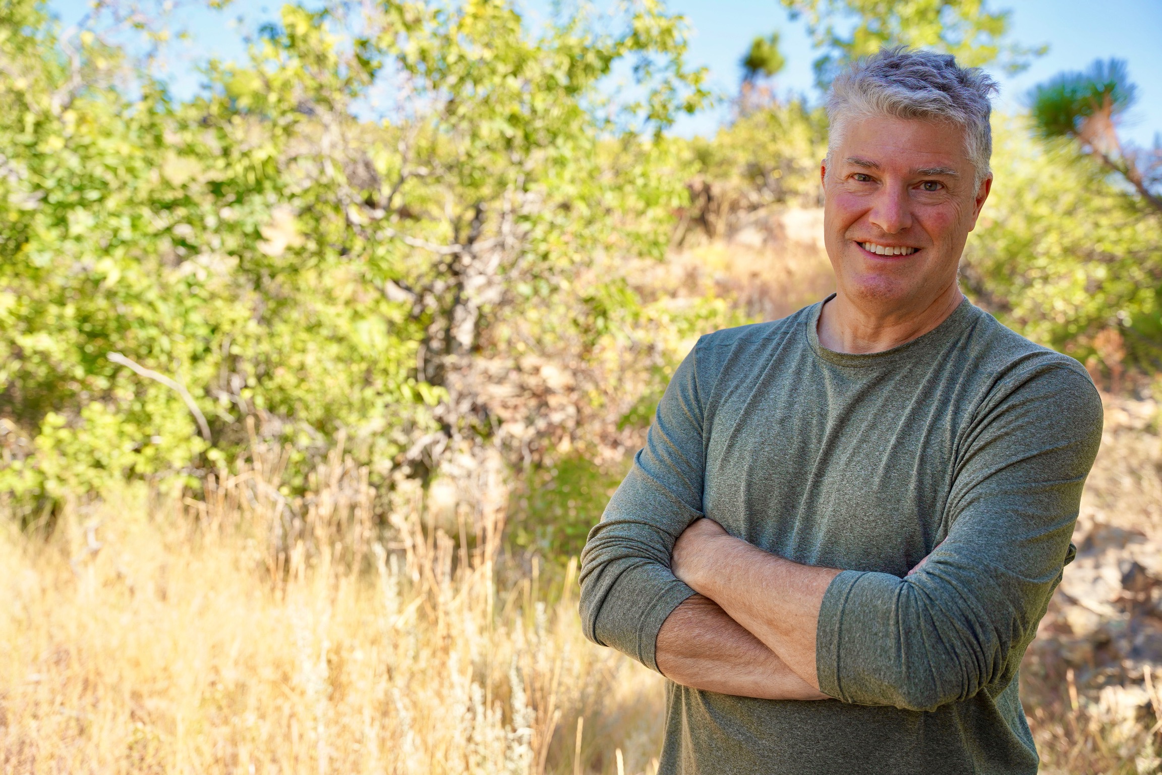 A confident, approachable man stands outdoors with arms crossed, surrounded by natural greenery and sunlight, representing a welcoming and experienced leadership coach at Whisper Ranch, the retreat location for The Leader Coach.