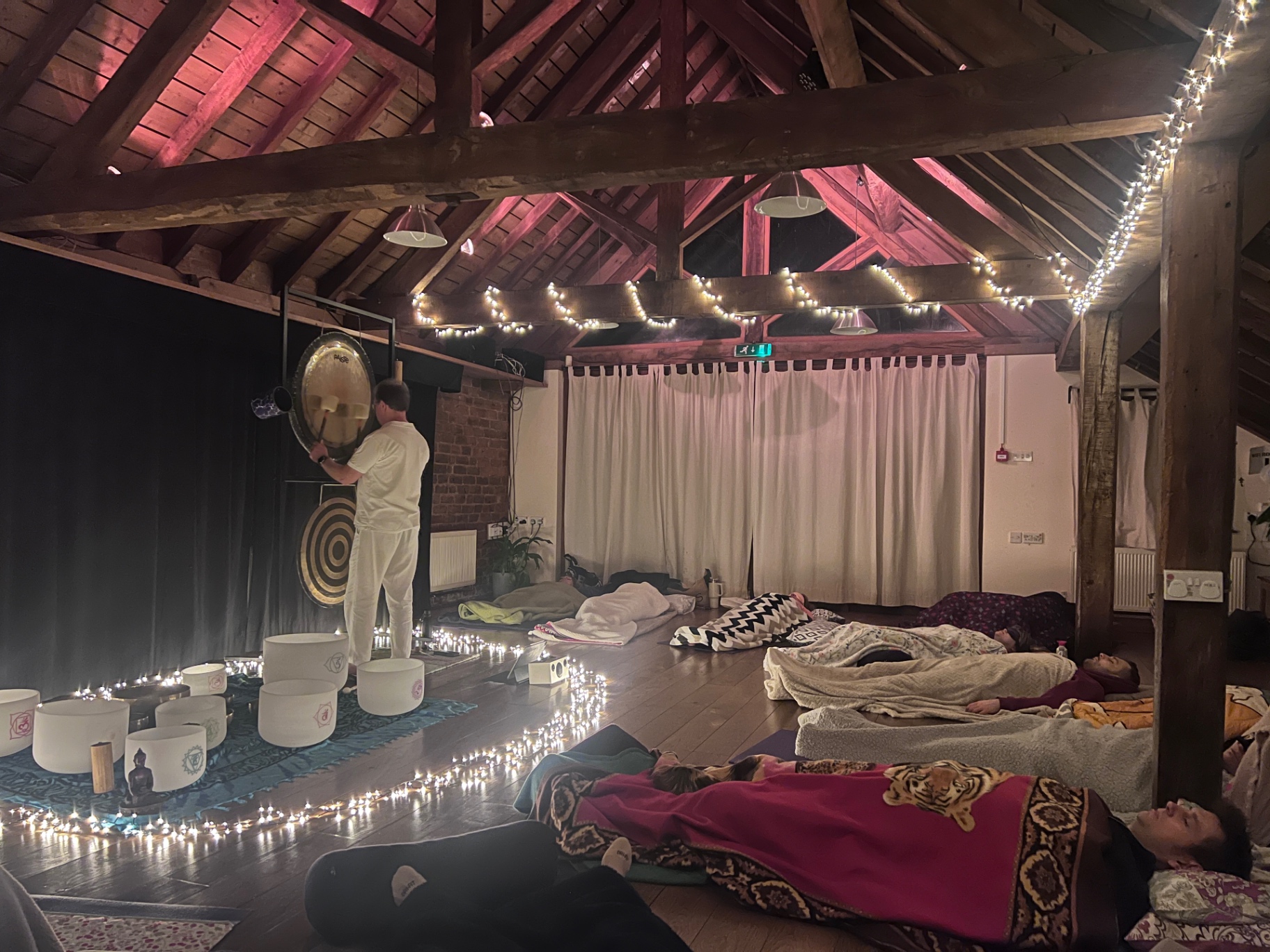 Sound Bath Meditation Session A tranquil sound bath meditation session in a softly lit studio. Participants lie under blankets on yoga mats while a gong practitioner stands surrounded by crystal singing bowls and gongs. Fairy lights outline the central sound area, creating a relaxing and peaceful atmosphere.