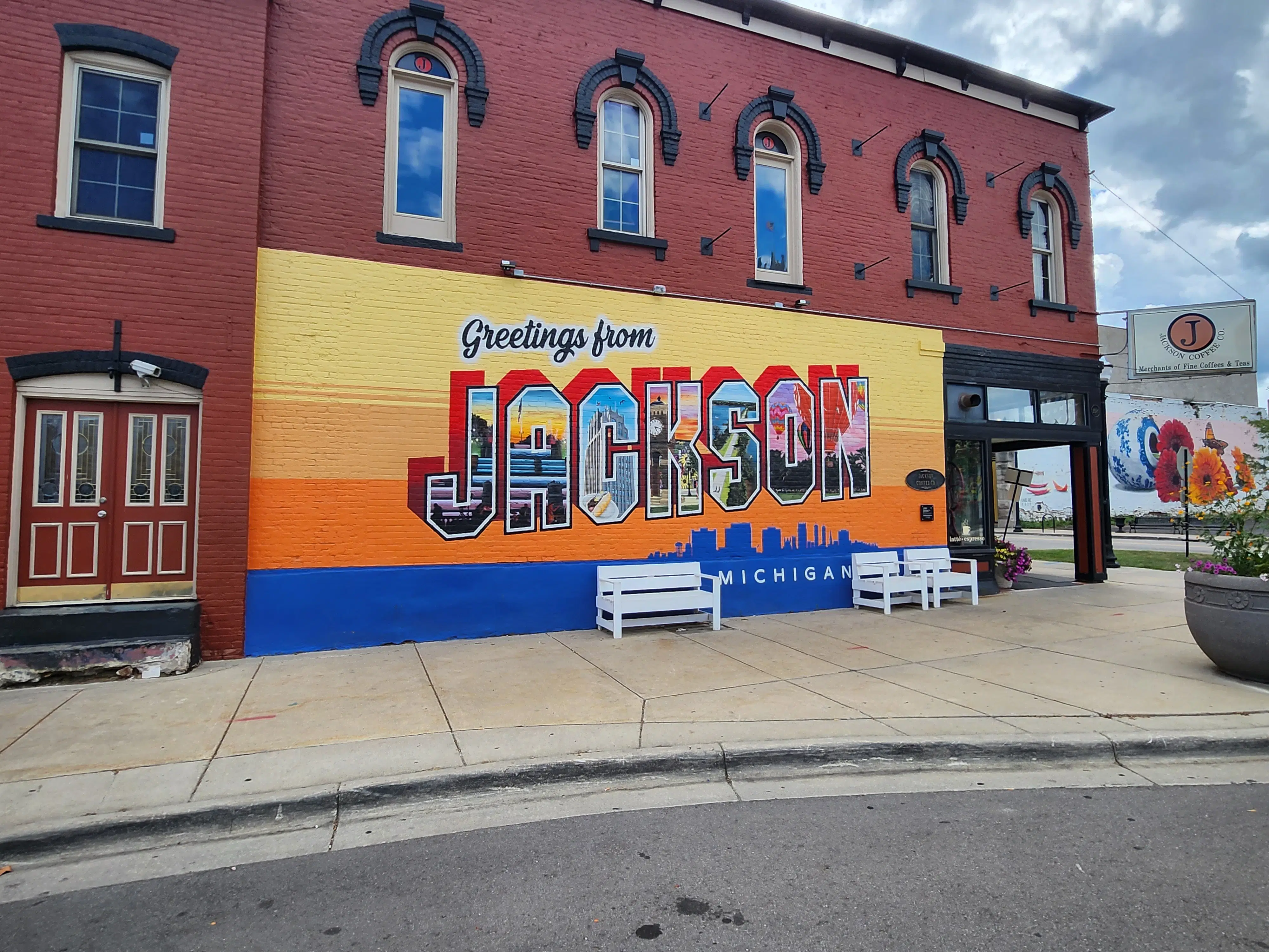 Colorful mural on a red brick building in downtown Jackson, Michigan featuring large text “Greetings from Jackson” and local landmark imagery.