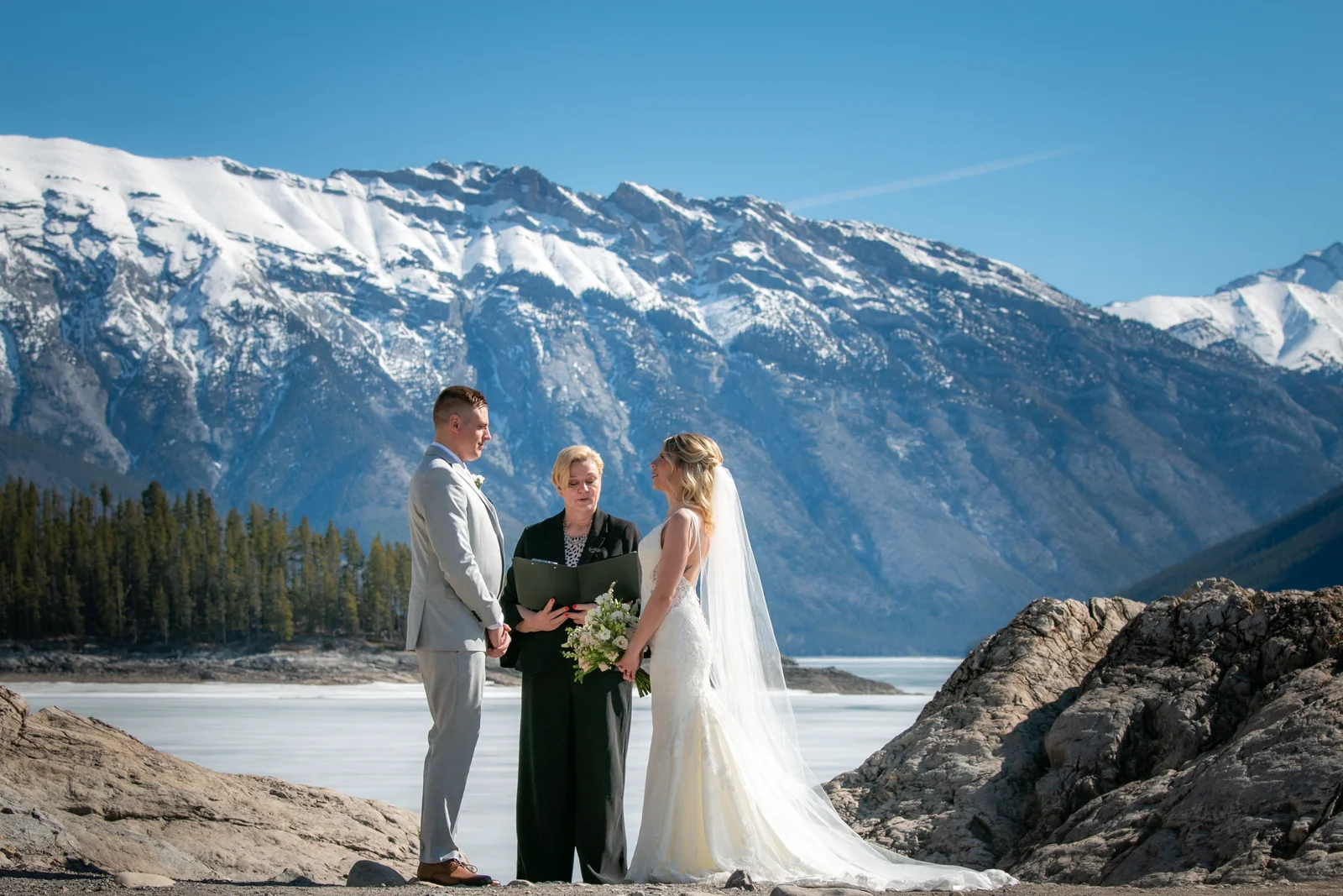A couple stands with an officiant on rocky terrain at the edge of a frozen lake, exchanging vows under clear blue skies.