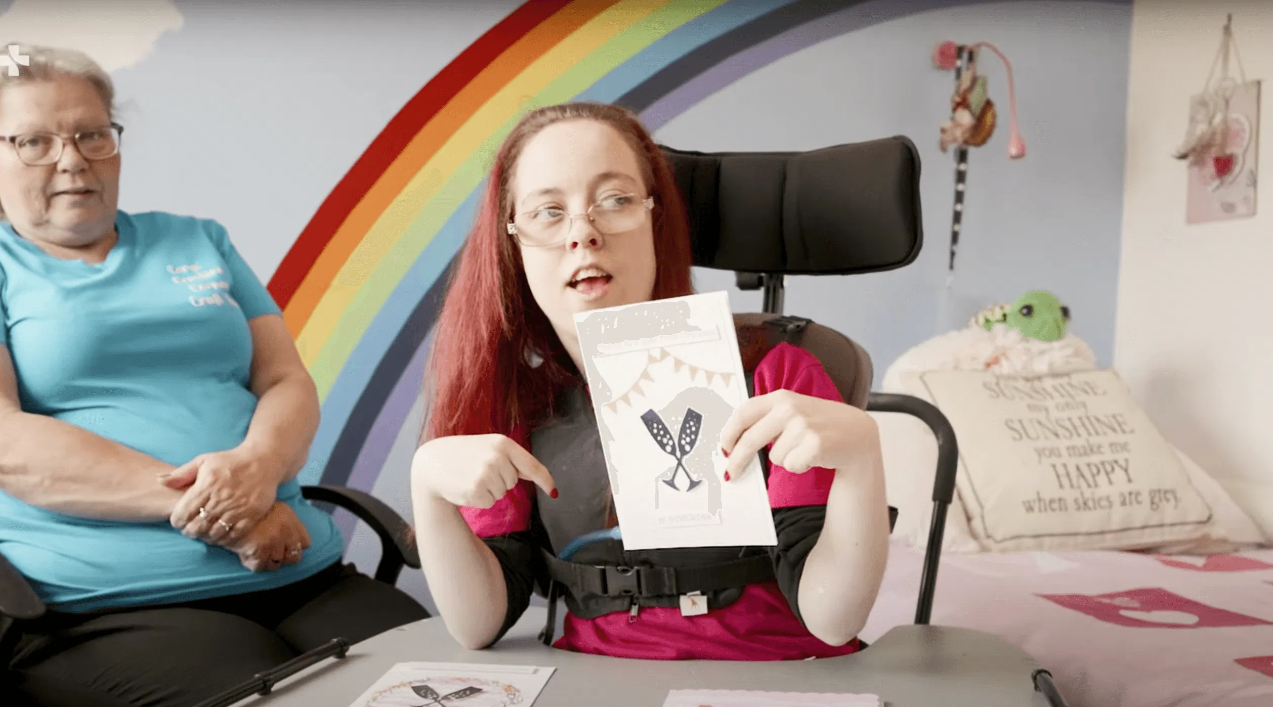A young woman in a wheelchair displays a handmade card during a creative home activity, supported by a carer, showcasing a positive, person-centred environment for independence.