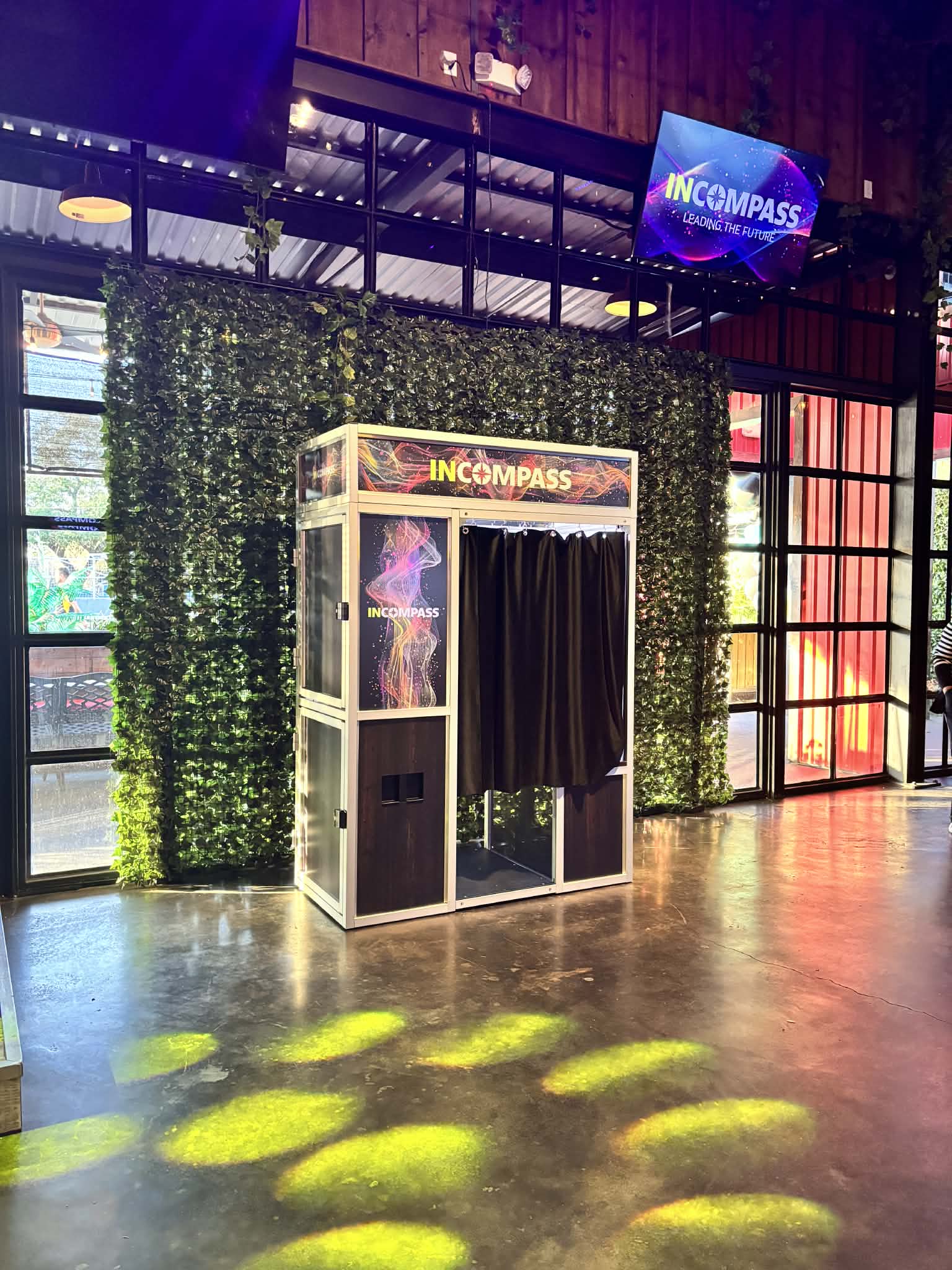 A branded modern photo booth setup for INCOMPASS, positioned indoors against a faux greenery wall