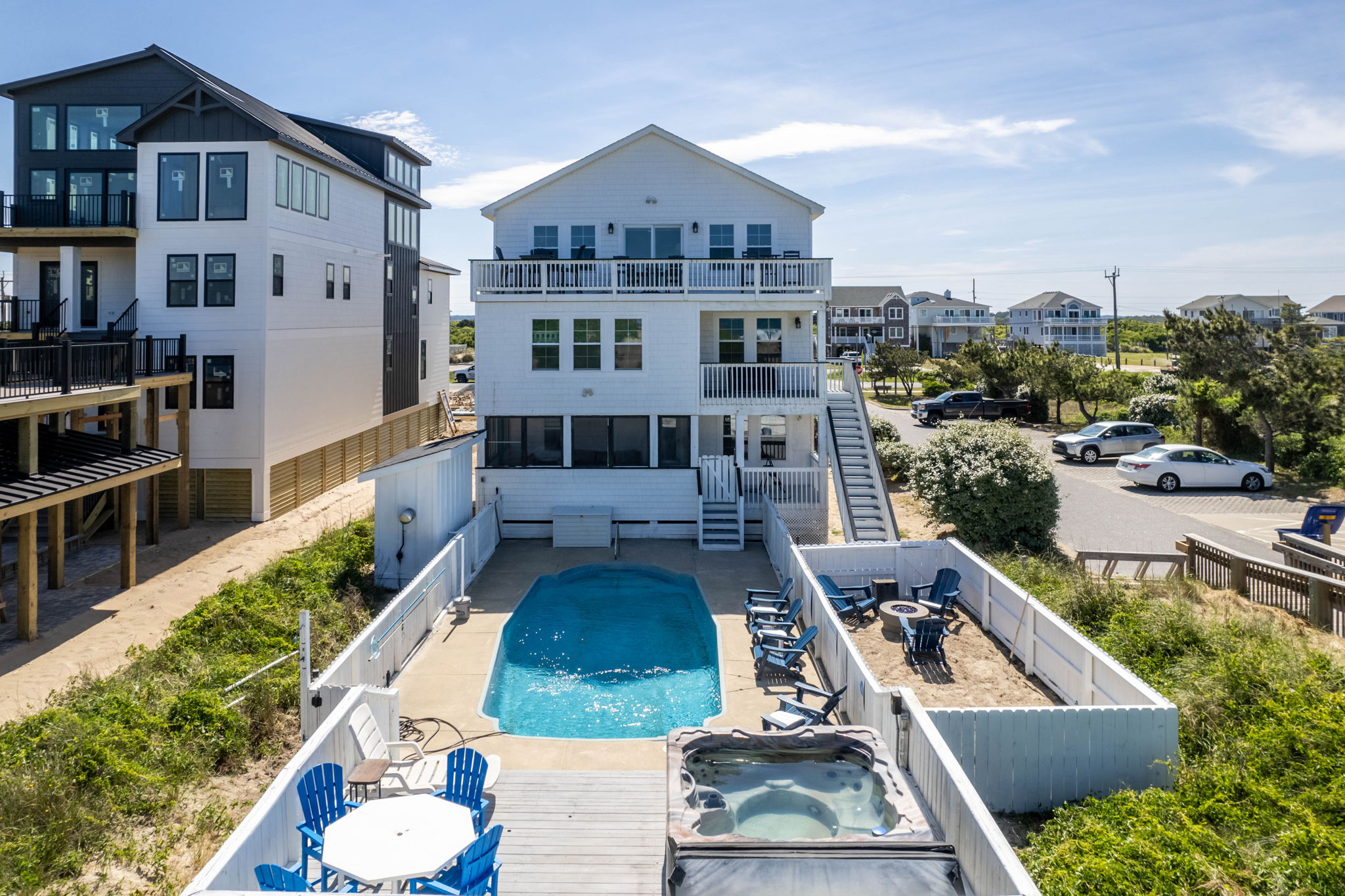 Three-story oceanfront luxury rental Luxury three-story oceanfront rental featuring a private pool, hot tub, multiple decks, fire pit area, ample outdoor seating, and fenced yard. Modern white exterior, next to beach access, and parking for several vehicles. Perfect for large groups or family gatherings.