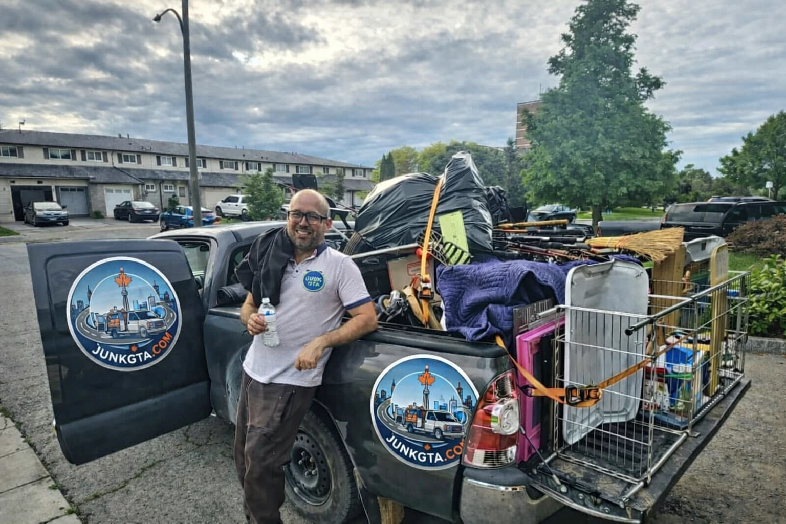 A Junk GTA crew member stands beside a pickup truck loaded with various junk items, such as large bags, a broom, a pet cage, miscellaneous furniture, and household goods