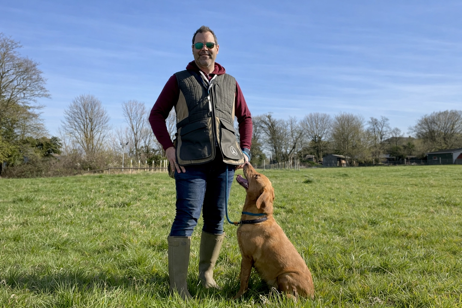 A gundog trainer stands in a grassy field with a focused gundog sitting calmly at their side, ready for a personalised 1 to 1 training session.