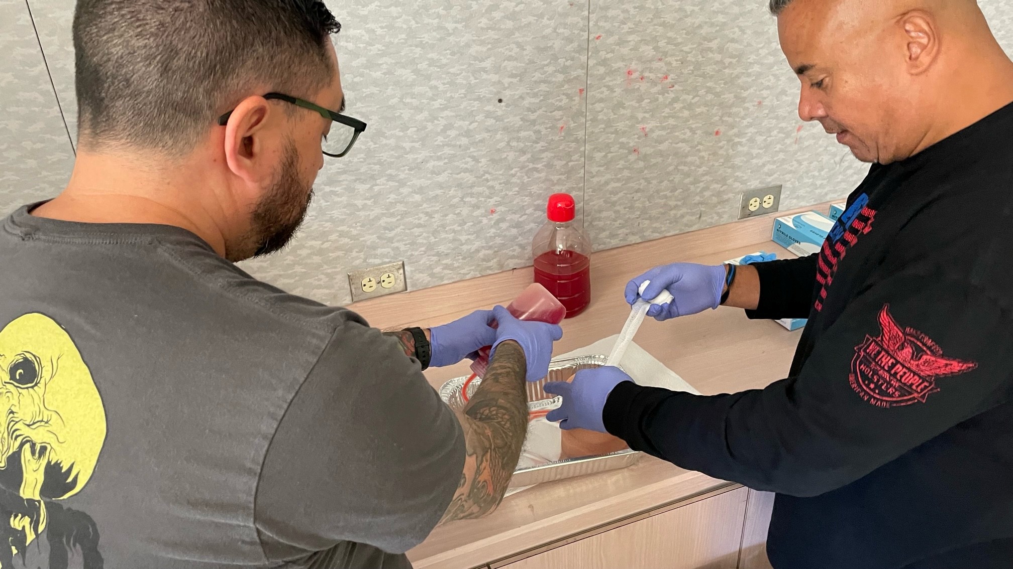 Two participants wearing gloves practice wound packing techniques using a training aid and simulated blood during a tactical first aid class.