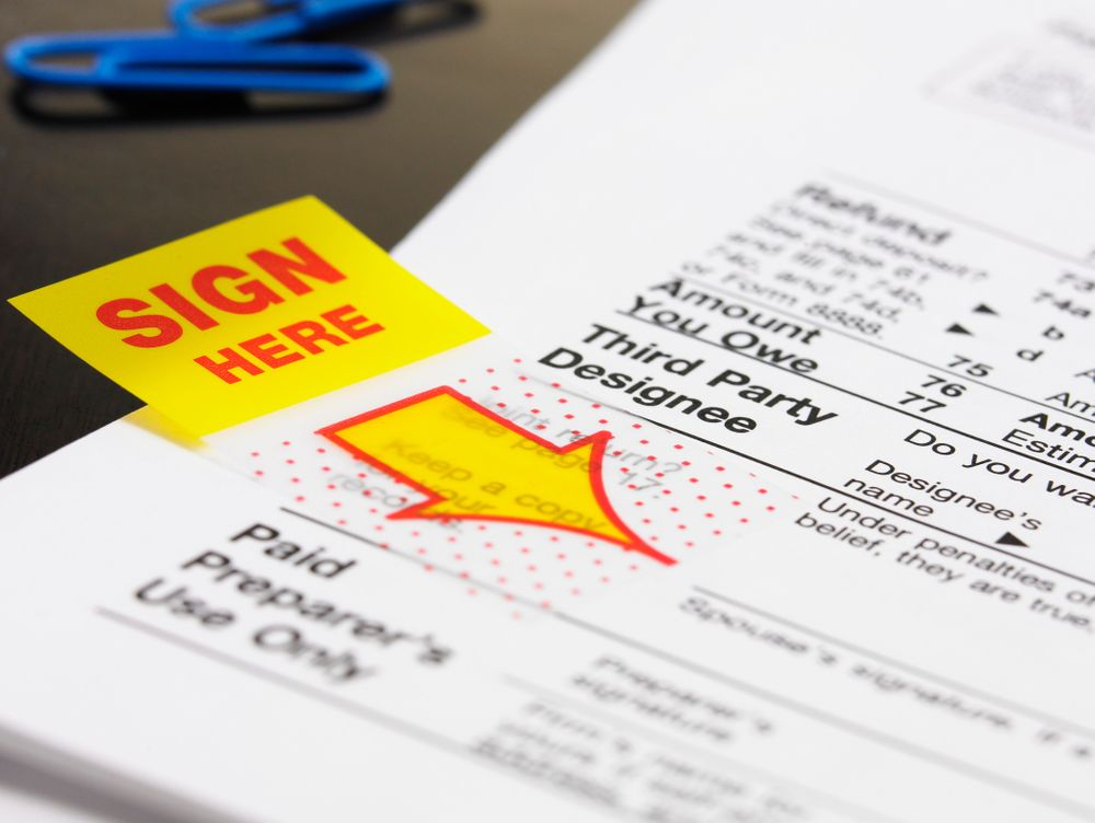 Tax Document Signature Section A close-up of a tax document with a yellow 'SIGN HERE' sticker attached, highlighting the signature section for the taxpayer. The form displays sections for third party designee and preparer's information, indicating a professionally prepared tax return ready for client review and signature at Brick Taxes LLC.
