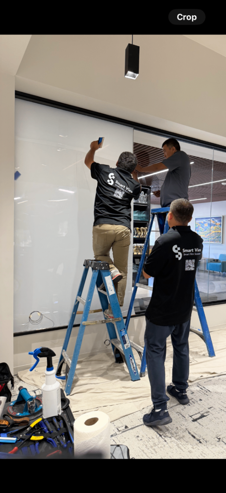 Smart View Smart Film Solutions Installation Smart View Smart Film Solutions installation team applying proprietary PDLC smart film to a large glass wall in a commercial interior. Technicians are using ladders and professional tools for precise, edge-to-edge fitting, ensuring seamless switchable privacy functionality. Branded uniforms emphasize company identity and expertise.
