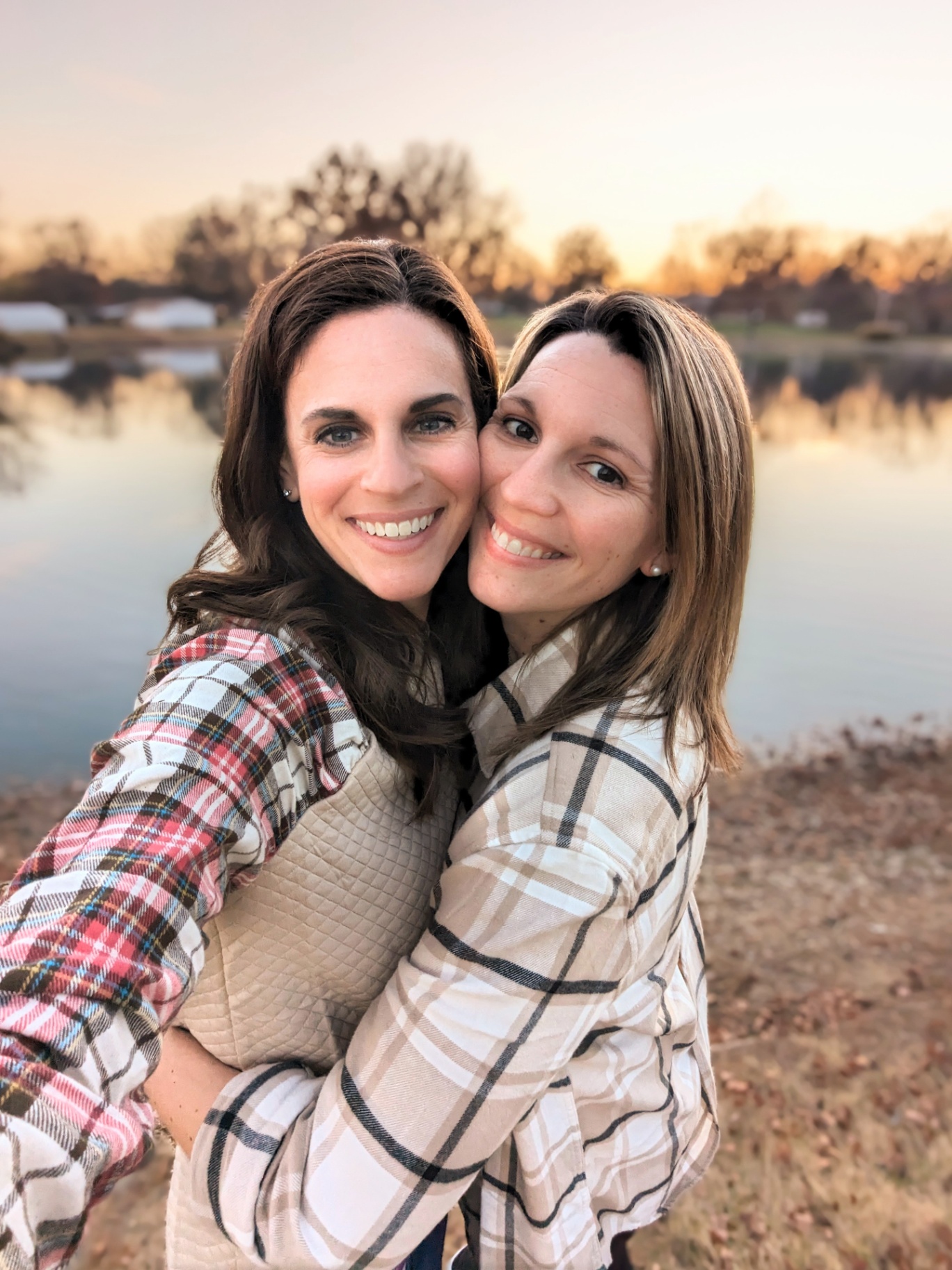 Two women stand close together outdoors by a lake at sunset, happily smiling and embracing.