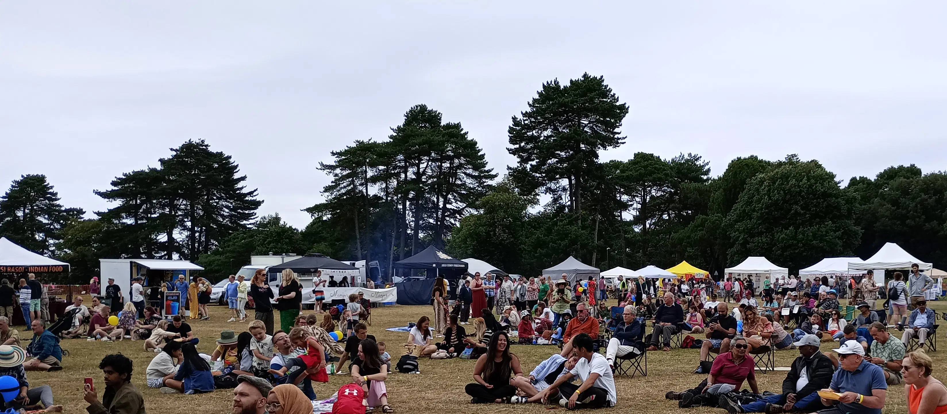 Families and friends gather on the grass at the Beautiful World Festival, enjoying live performances and diverse global street food from colorful market tents and food stalls.