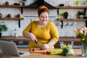 A woman prepares fresh vegetables while referencing a laptop in a bright, modern kitchen.