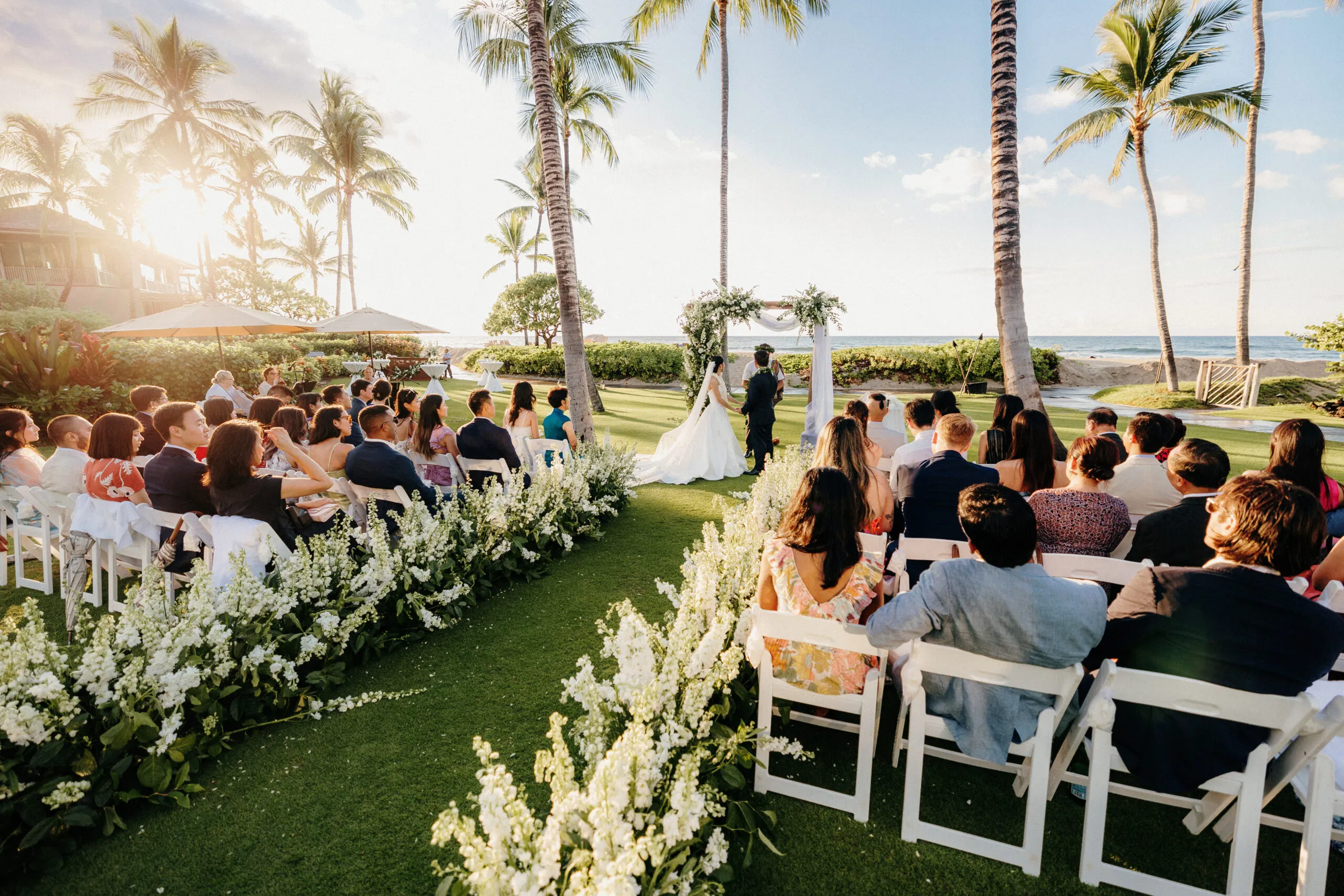 An outdoor wedding ceremony on the Big Island, set on lush green grass with ocean views and a floral arch.