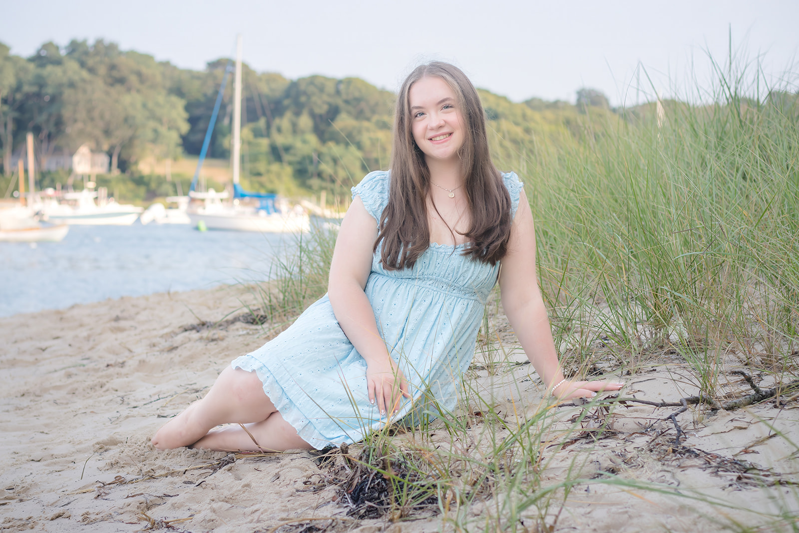 Smiling woman in a light blue dress sits on sandy beach, surrounded by grass. Boats and trees are visible in the background during her Falmouth Senior Portrait