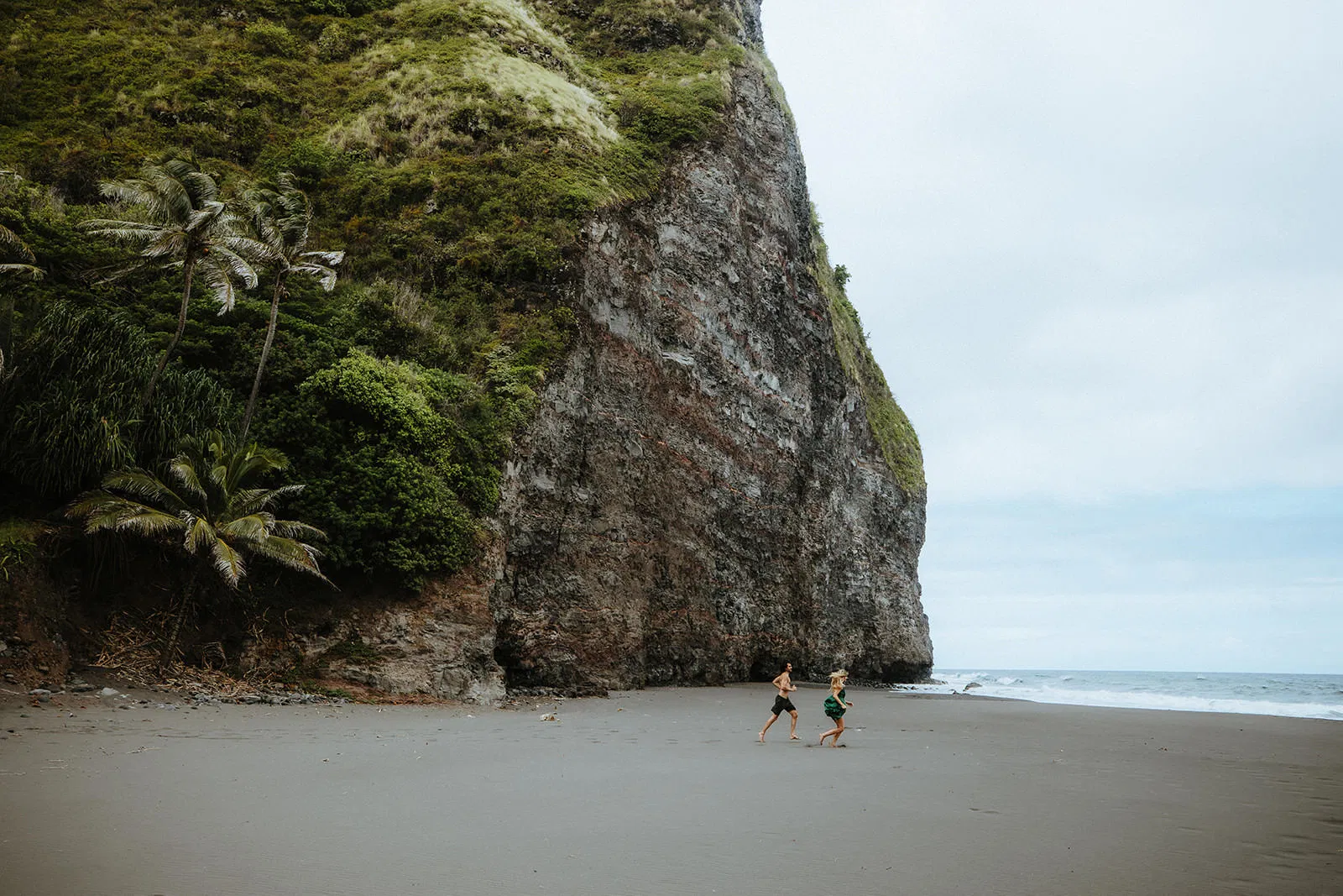 A playful couple runs barefoot along a black sand beach at the base of a dramatic green cliff.