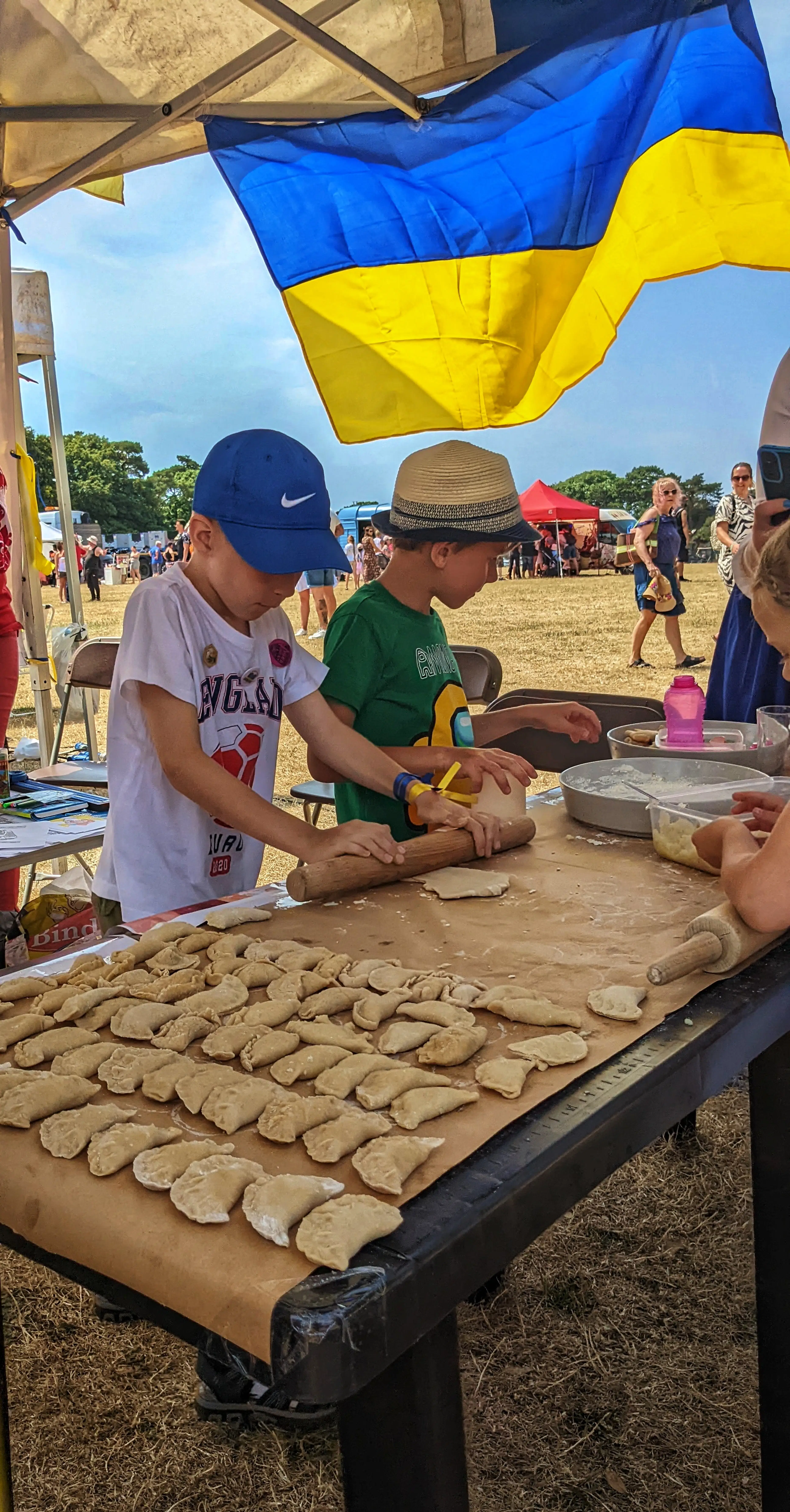 Children participate in a hands-on Ukrainian dumpling making workshop under a tent, guided by adults.