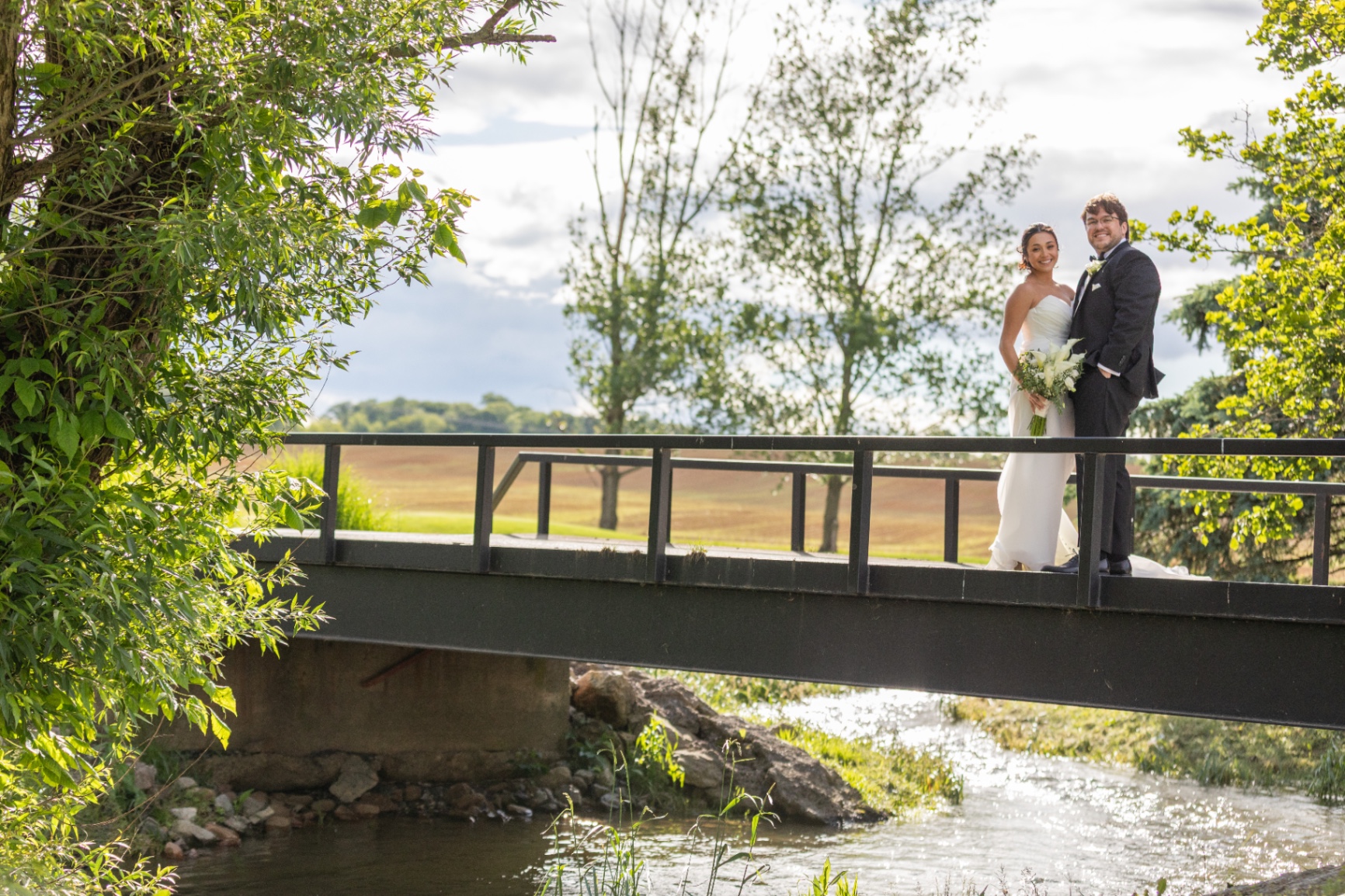 A bride and groom stand together on a small bridge over a sunlit creek, smiling softly