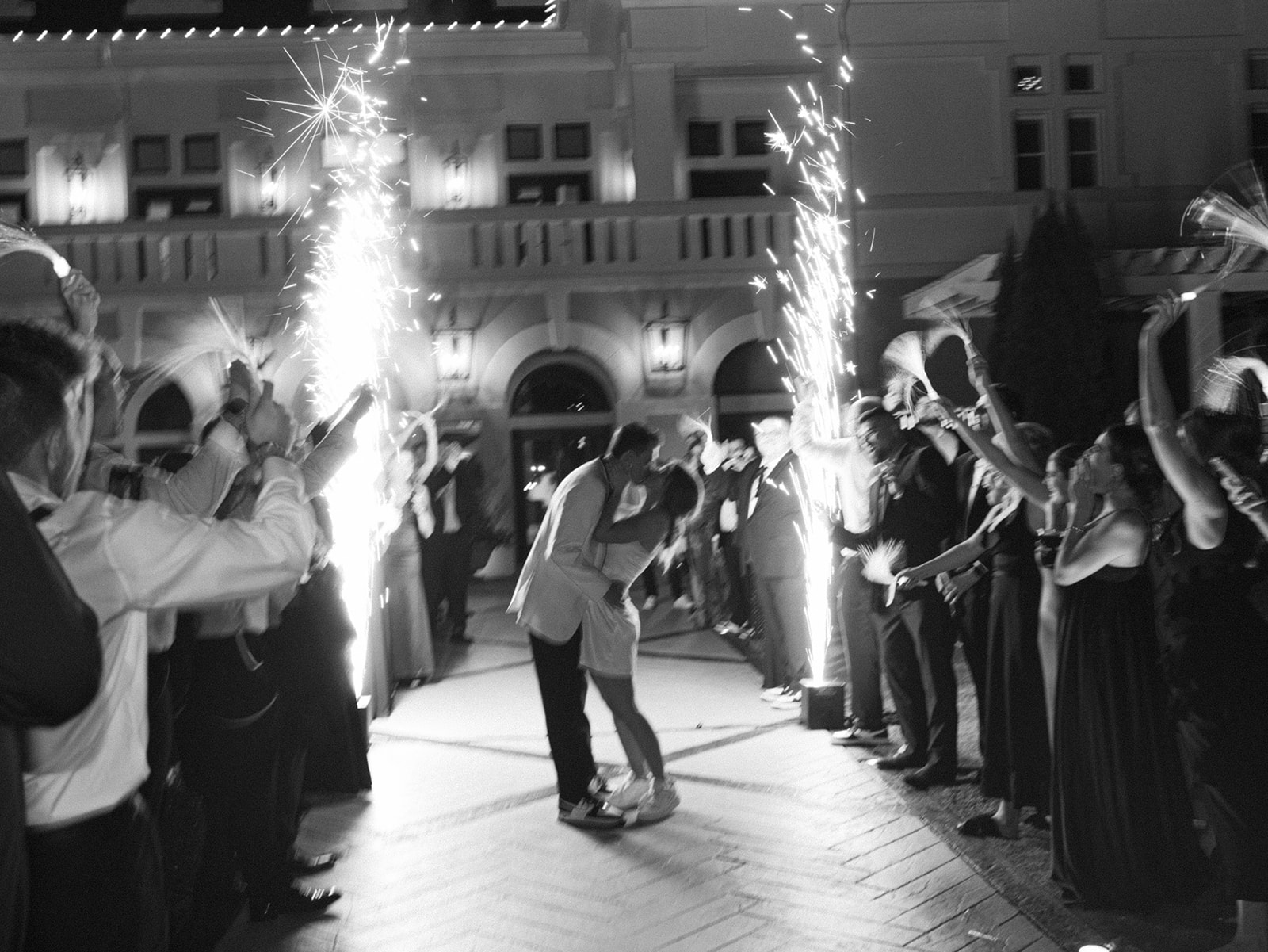 Wedding Exit Kiss A wedding couple shares a kiss during their grand exit, surrounded by cheering guests holding sparklers and cold spark fountains. JAMMIN' DJs' custom event lighting and atmospheric effects create a dramatic and festive atmosphere.