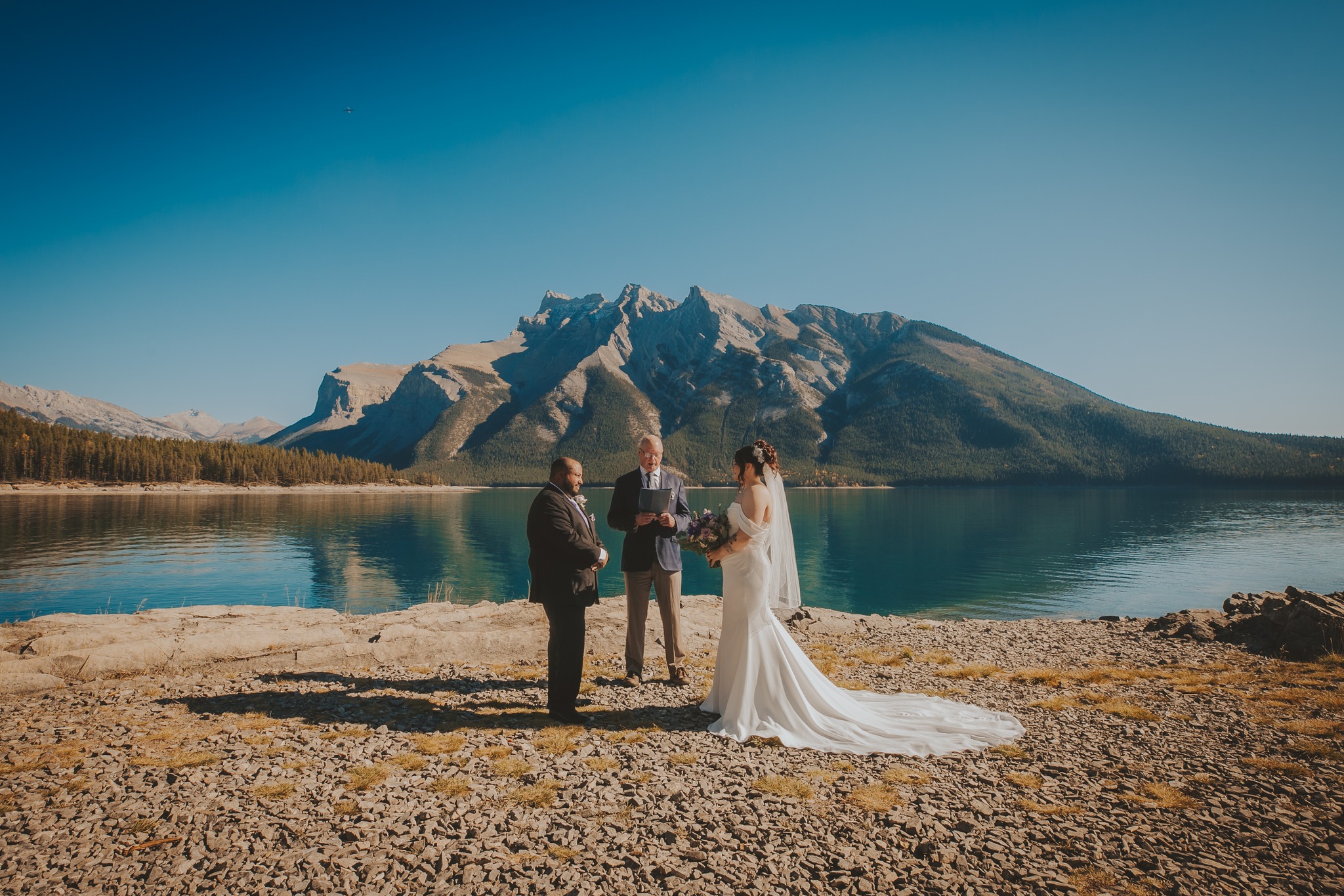 A couple exchanges vows with an officiant on the rocky shore of Lake Minnewanka under clear blue skies.