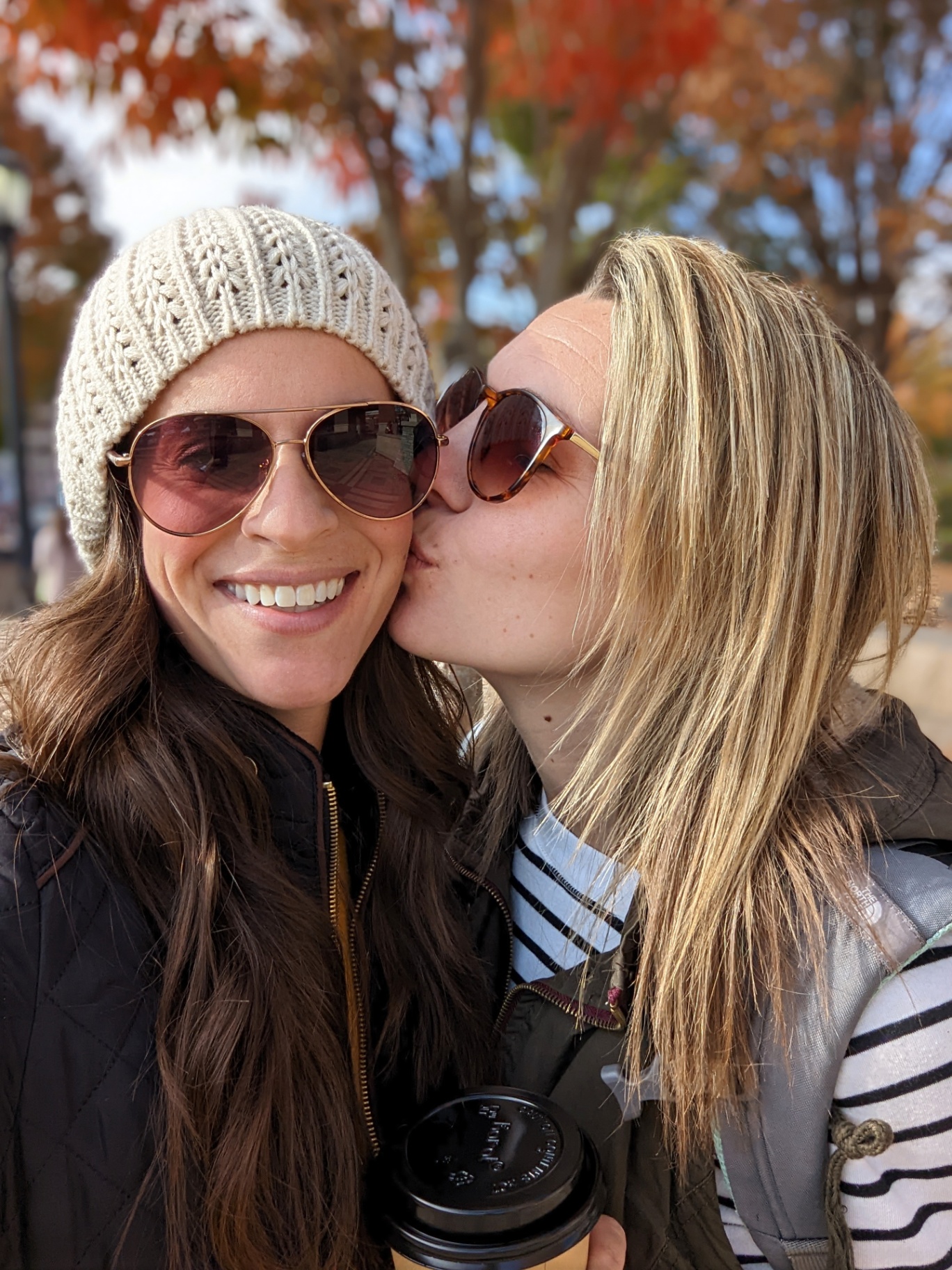 Two women smiling closely together outdoors, one kissing the other on the cheek