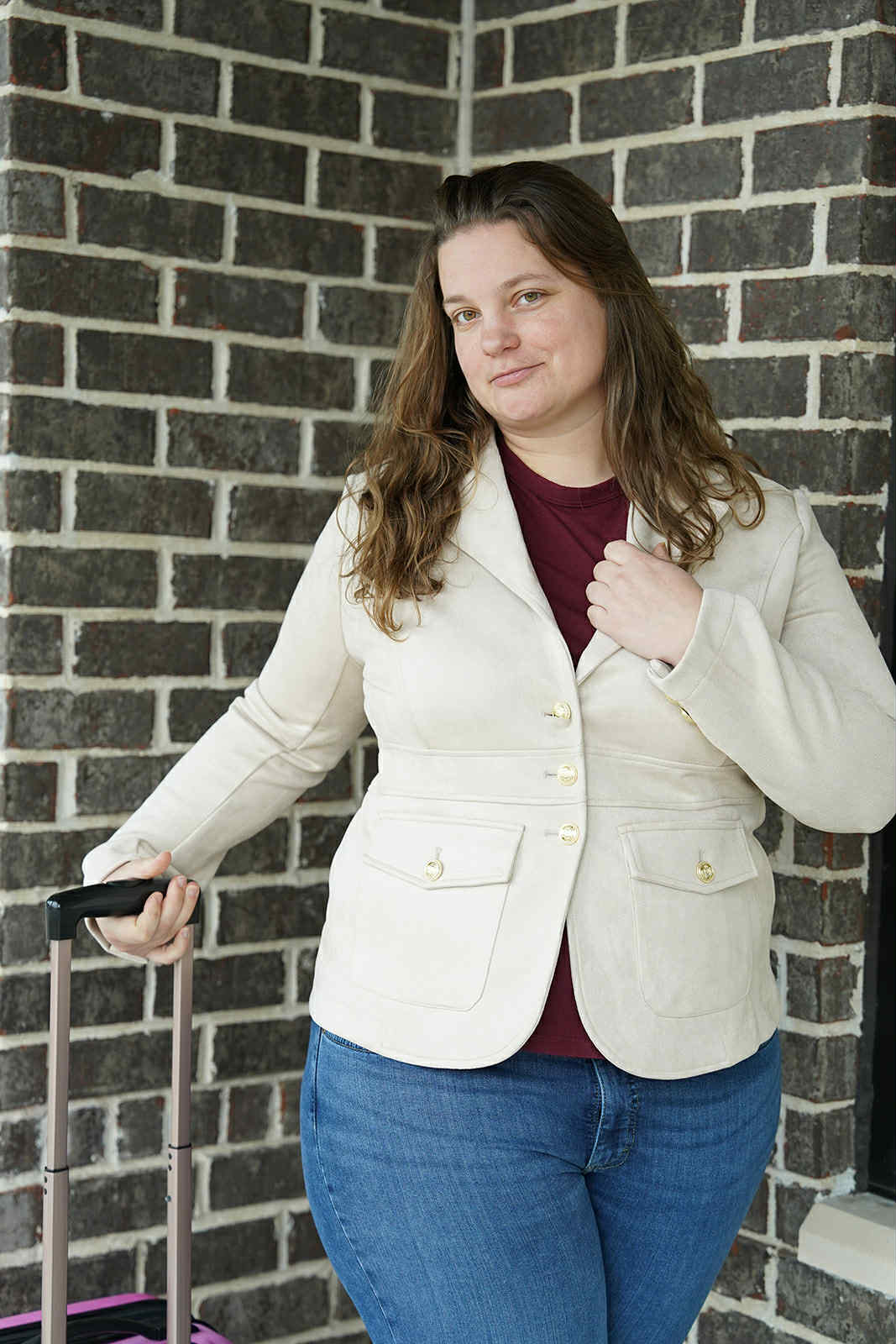 Woman ready for premium travel A woman stands outside near a brick wall, dressed in a cream blazer and jeans, holding the handle of a suitcase, ready to embark on a premium, well-organized travel experience arranged by Freedom to Explore Travel Agency.
