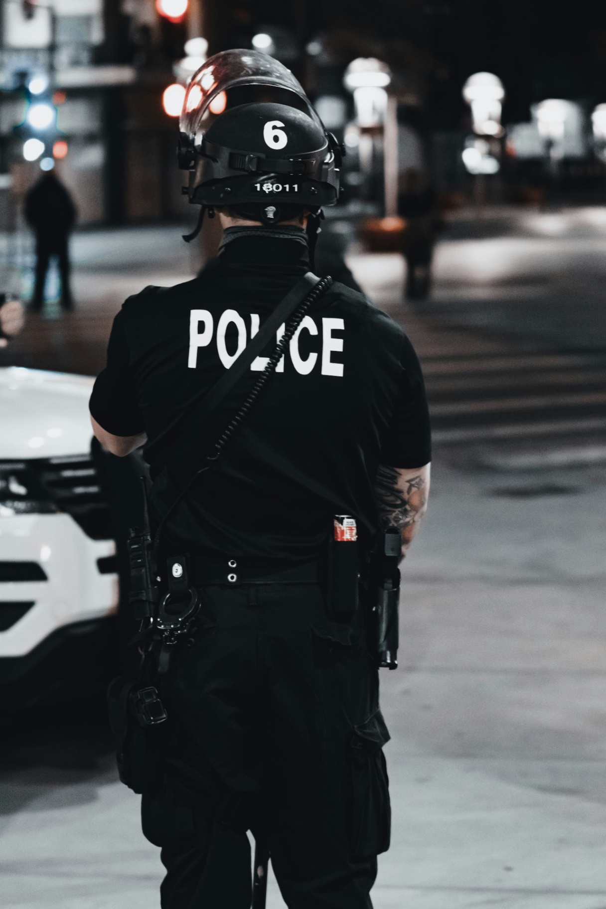 A police officer in full uniform and tactical gear stands near a patrol vehicle at night