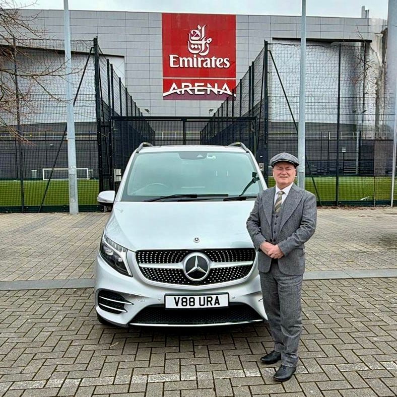 A professional chauffeur in a tailored suit stands beside a luxury silver Mercedes-Benz V-Class with Aura Journeys branding at Emirates Arena.