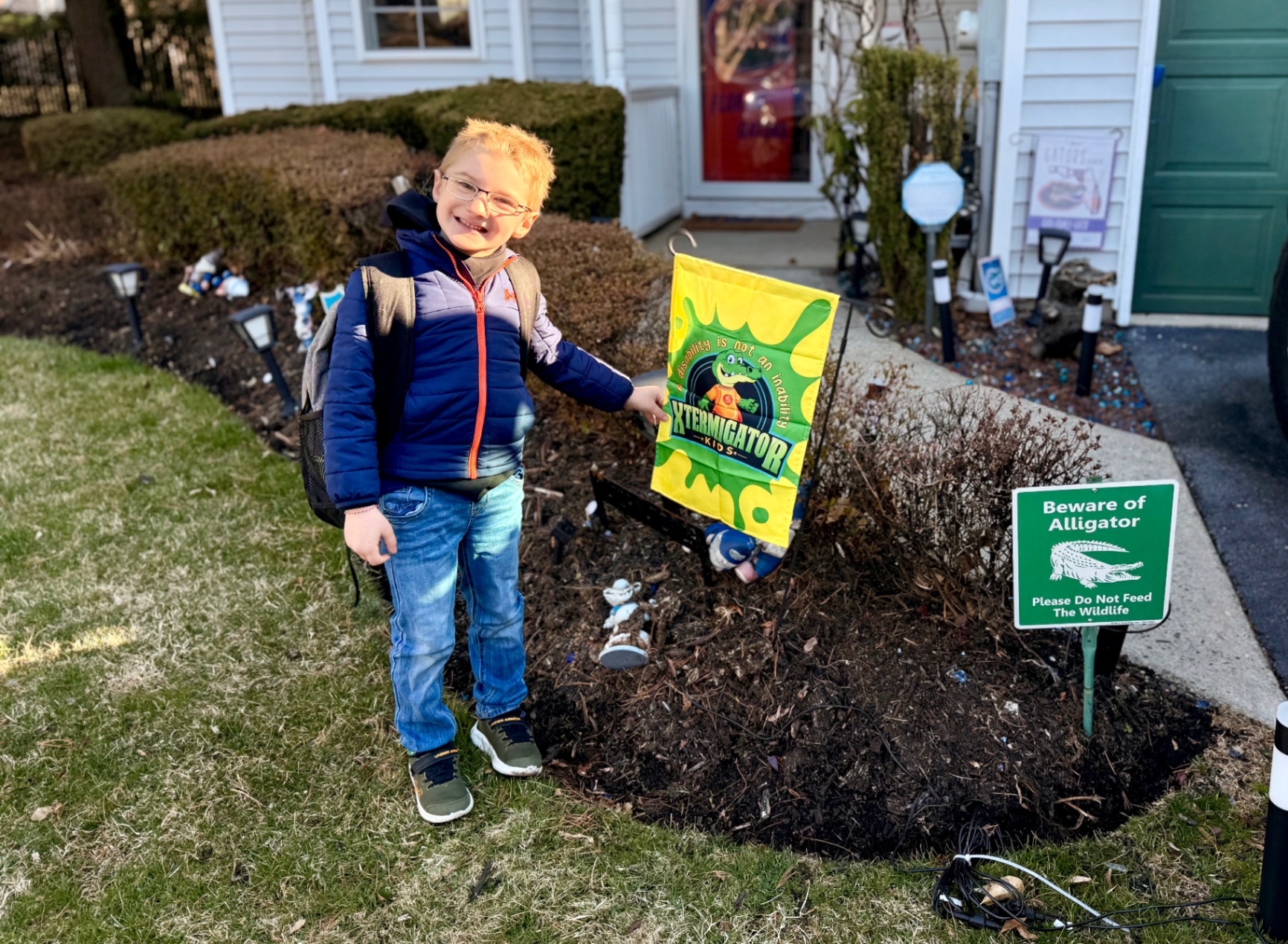 XTERMIGATOR KIDS Inclusive Flag Photo A smiling child with a backpack stands outside a home, holding a bright XTERMIGATOR KIDS flag featuring the Friendly Ferns Swamp character and the motto 'A disability is not an inability.' A green 'Beware of Alligator' sign is posted in the garden, promoting the brand's playful, inclusive, and educational messaging about disabilities.