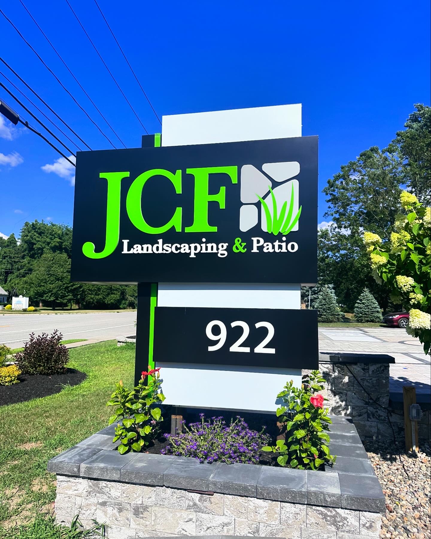 Custom monument sign for JCF Landscaping & Patio A custom monument sign featuring bold green and white channel letters and address numbers on a modern black and white background. The sign is installed on a stone base with an integrated planter, showcasing craftsmanship and full-service installation for enhanced business visibility.