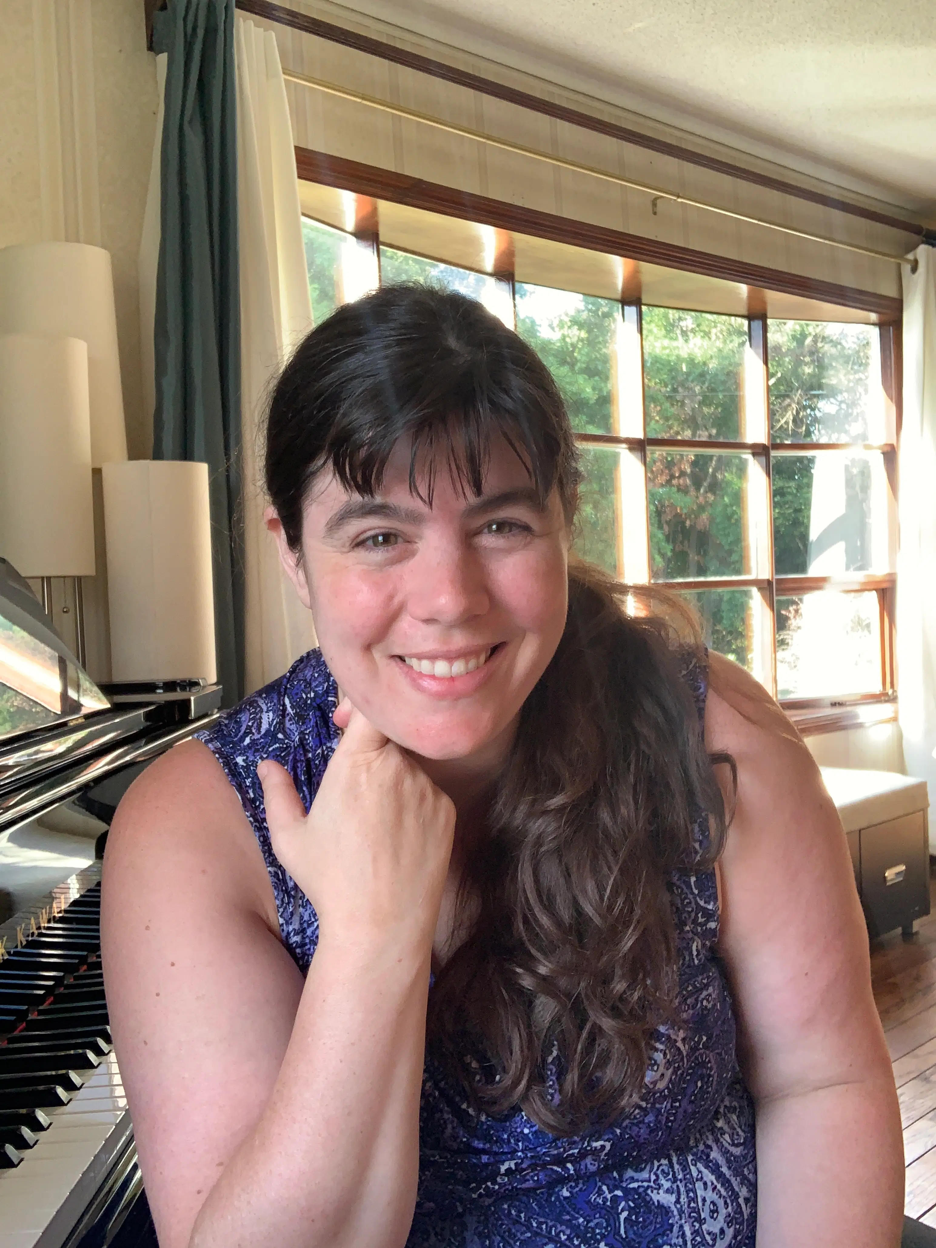 A smiling woman sits beside a grand piano in a sunlit music studio, embodying the welcoming environment