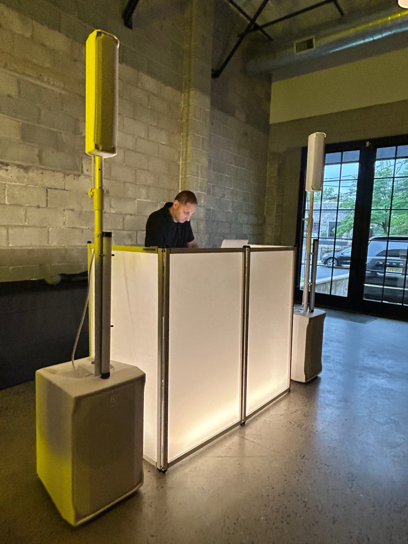 Modish Entertainment DJ at Upscale Industrial Event A Modish Entertainment DJ stands behind an illuminated modern DJ booth, flanked by two high-end column speakers, preparing for an upscale event in an industrial-style venue with natural light and exposed brick walls.