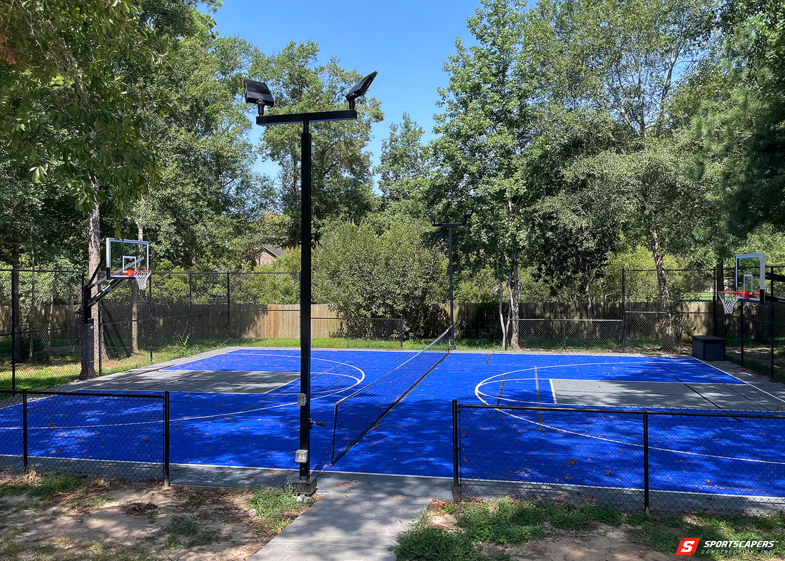 Full-sized outdoor multi-sport court featuring a blue playing surface, basketball hoops, and lighting poles for nighttime play.