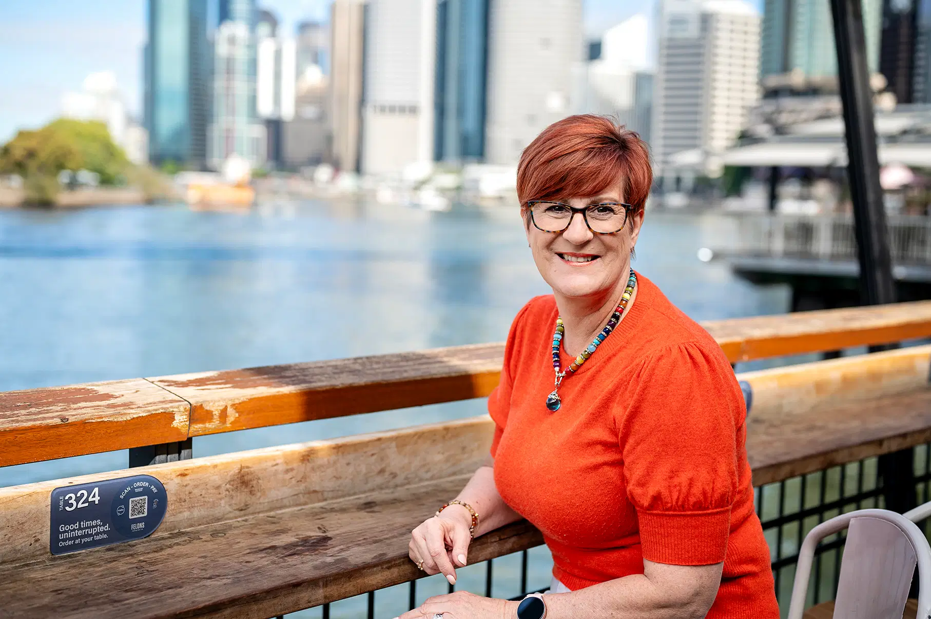 A woman dressed in a vibrant orange top sits at an outdoor waterfront café with a calming cityscape in the background, representing a relaxed yet professional approach to personalized travel planning.