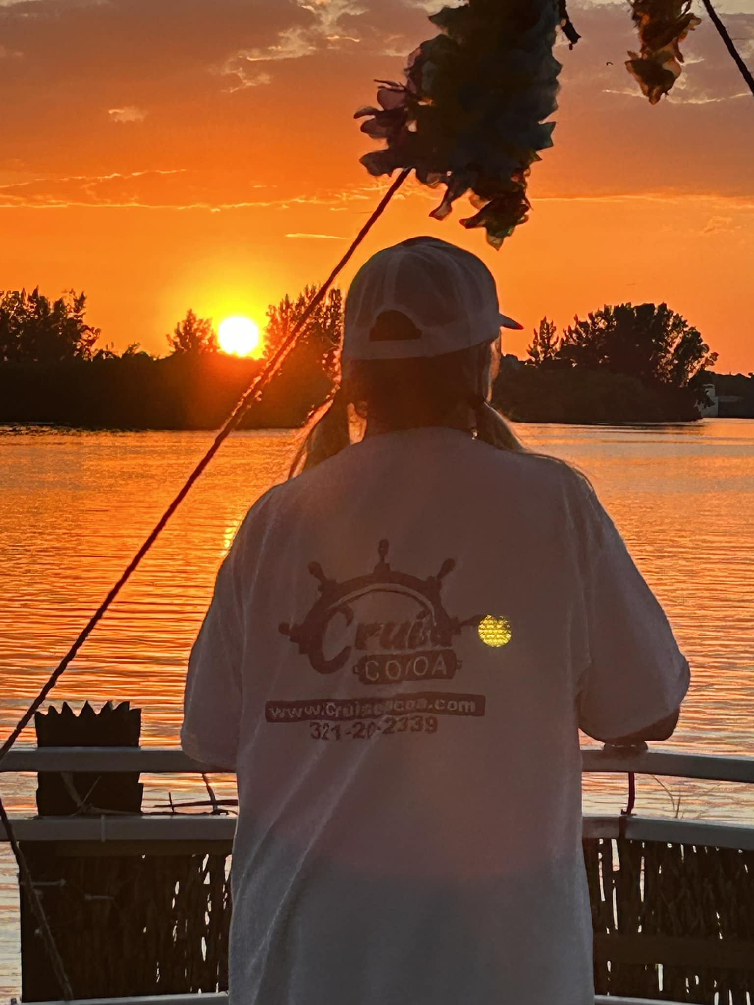 Cruise Cocoa Sunset Boat Crew A Cruise Cocoa crew member stands at the rail of a tiki-themed boat, wearing a branded shirt, watching a vibrant orange sunset over the Indian River Lagoon. The boat decor includes tropical garland, and calm waters reflect the sunset. Trees line the distant shore, creating a peaceful evening cruise atmosphere.