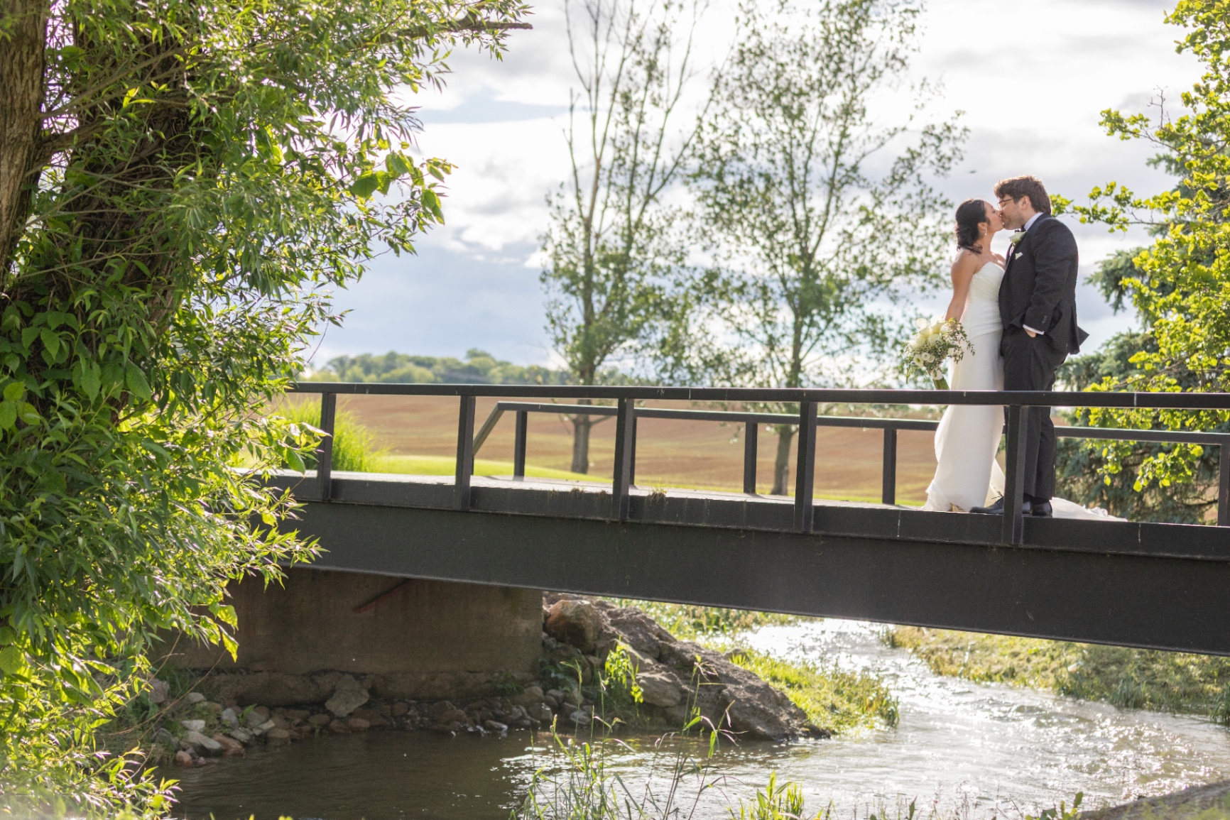 A bride and groom share an intimate kiss on a sunlit bridge over a gentle stream