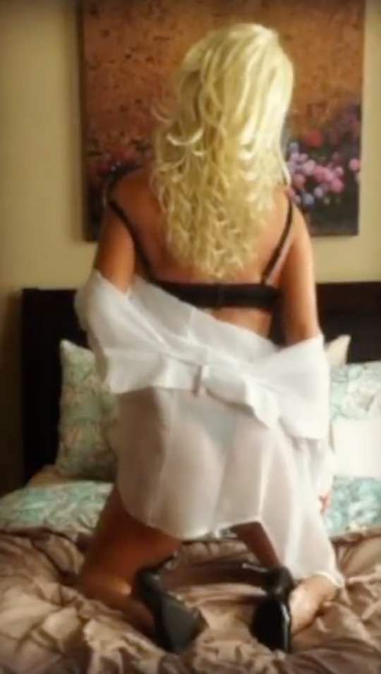 Maddie 31 A person with long, wavy blonde hair kneels on a bed, seen from behind, wearing a black bra, sheer white robe, and black high heels. The soft and inviting bedroom setting captures an intimate moment of self-expression