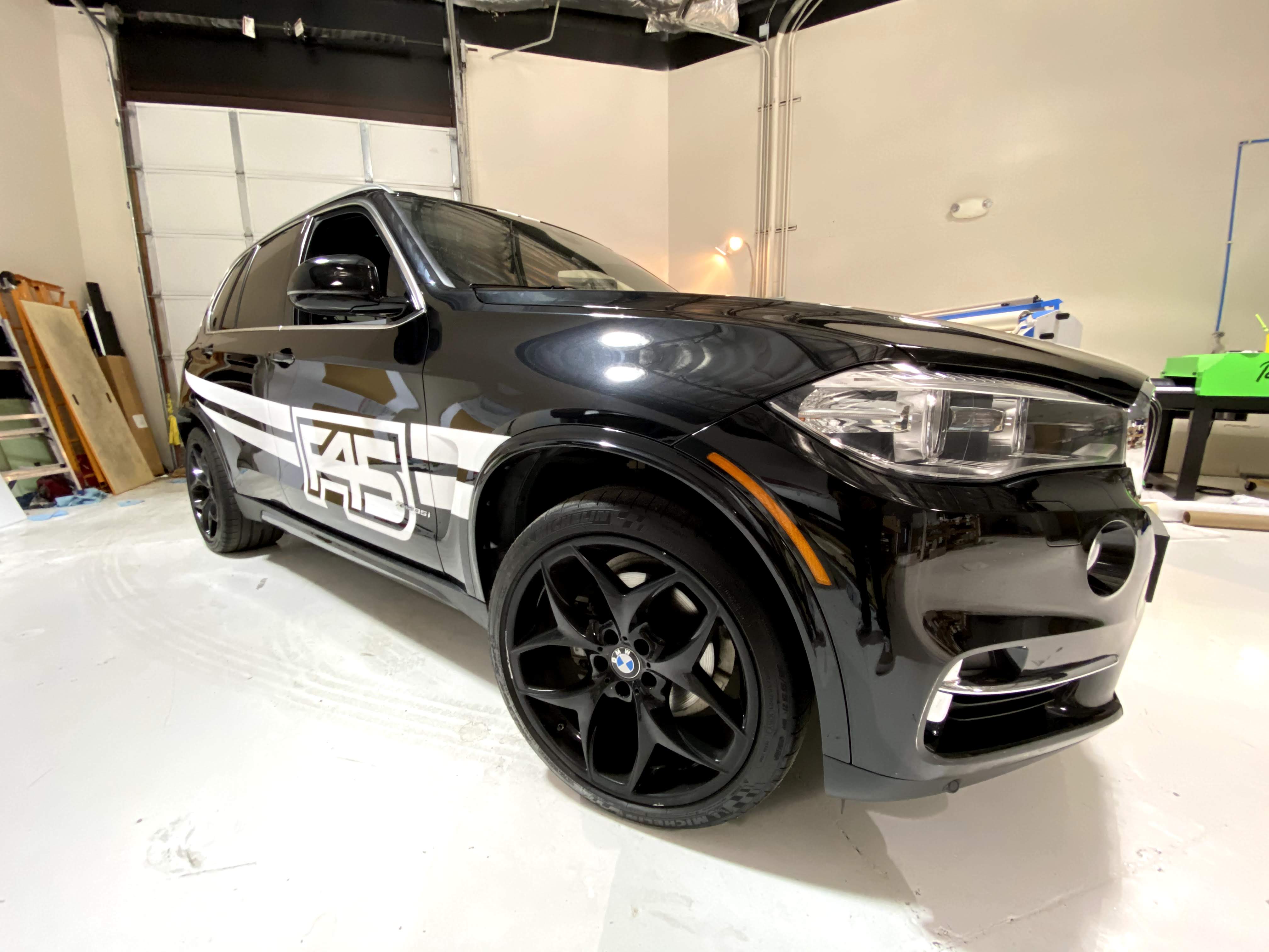 Black BMW SUV with FAB Logo Wrap Black BMW SUV featuring a high-impact custom vehicle wrap with bold white stripes and a large stylized 'FAB' logo on the front door, designed and installed in a professional garage environment to enhance mobile brand visibility.