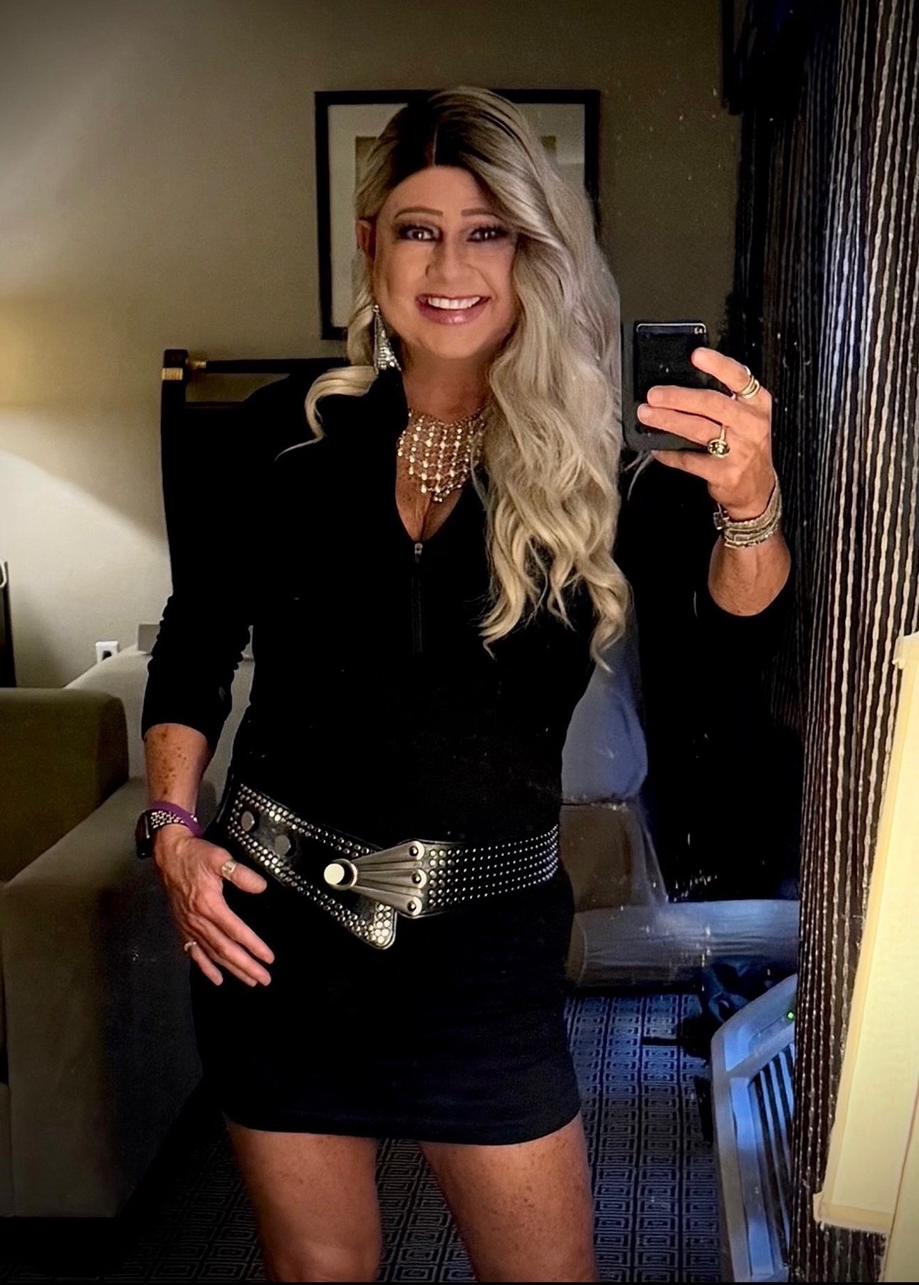 Maddie 21 A smiling transgender woman with long blonde hair takes a mirror selfie in a warmly lit room, wearing a black top, mini skirt, statement necklace, large earrings, and a studded belt. This image reflects the confidence, style, and authenticity celebrated in My Girl Life Podcast’s community.