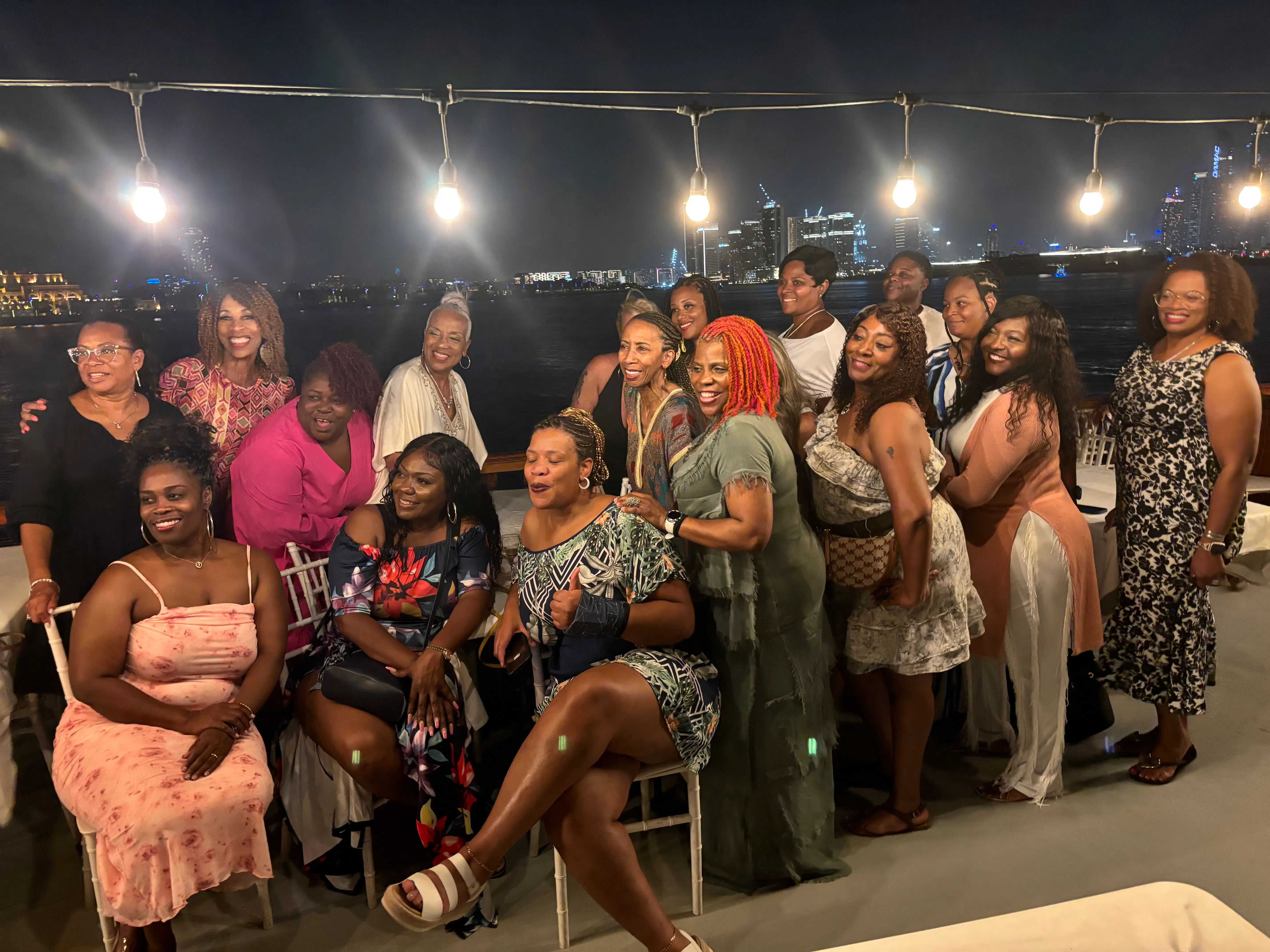 A joyful group of women poses together on an outdoor terrace at night, with the city skyline illuminated in the background.